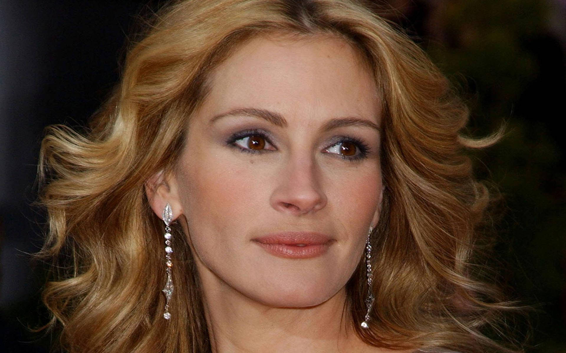 Julia Roberts 桌布 壁紙 — 一個女人的特寫肖像，她有波浪形金髮和閃爍的吊墜耳環，背景是黑暗模糊的。