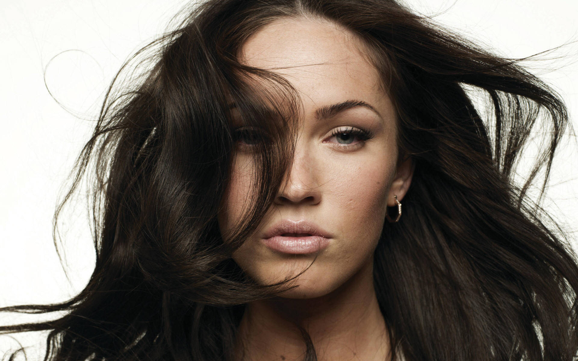Megan Fox HD 桌布 壁紙 — 一個女人的特寫肖像，她有深色的頭髮被風吹亂，眼神強烈，背景是純白色。
