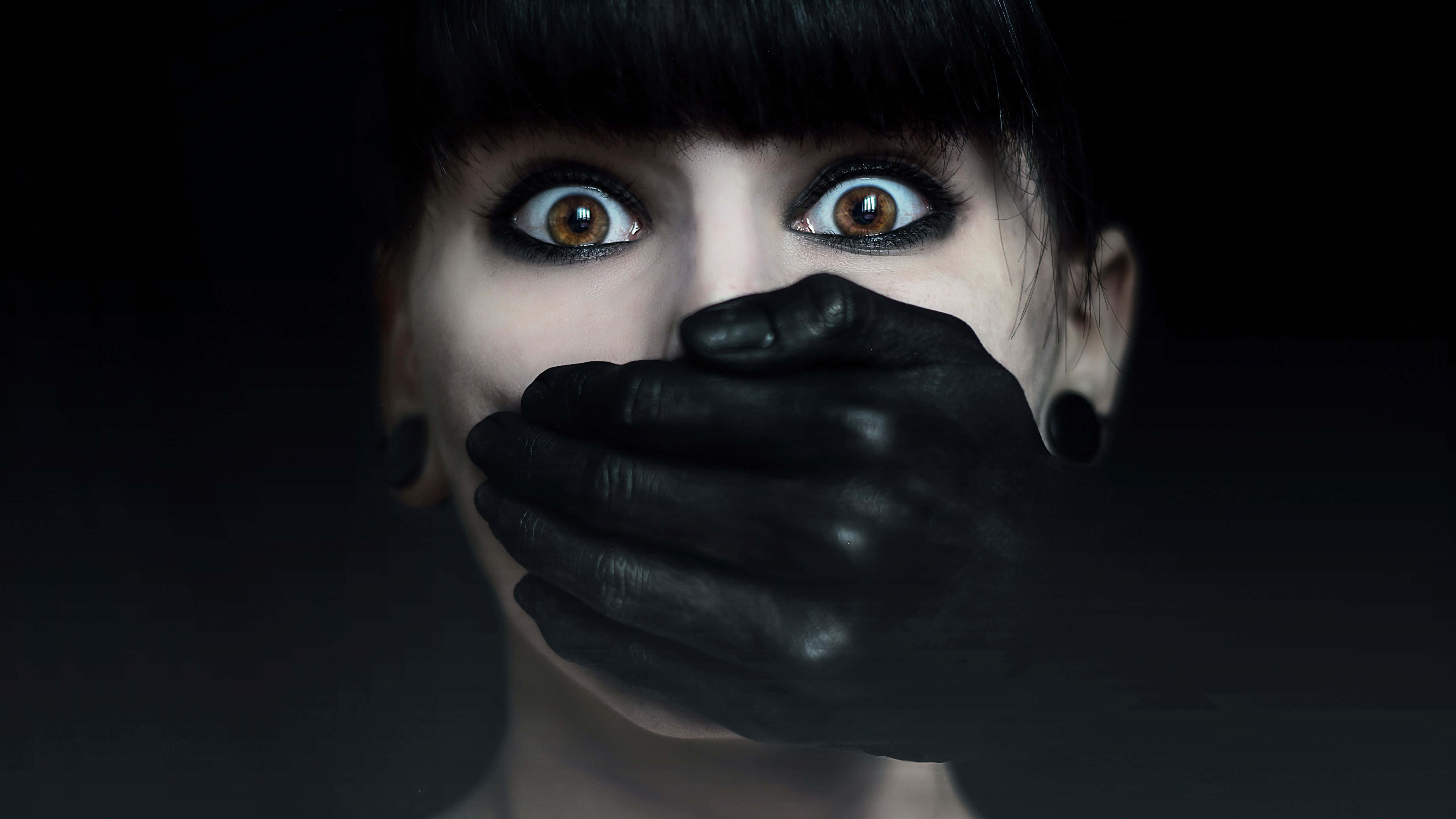 Dark Hand 桌布 壁紙 — 一個女人睜大驚恐的眼睛，凝視著一隻戴著手套的手在黑暗背景下捂住她的嘴。