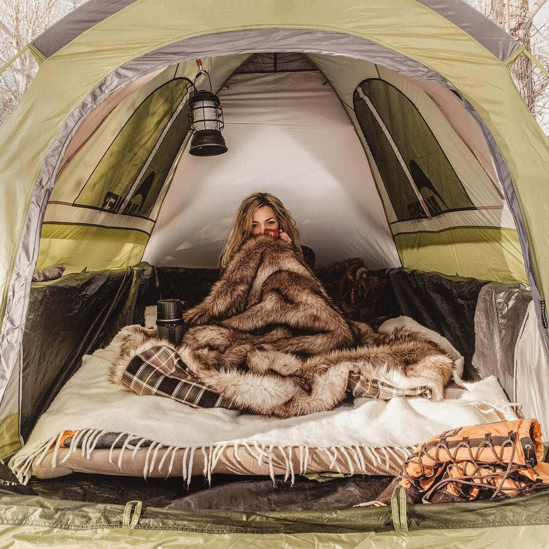 Winter Camping 桌布 壁紙 — 一個女人舒適地裹著毛茸茸的毛毯，坐在綠色露營帳篷內，在掛著的燈籠下啜飲杯子裡的飲料。