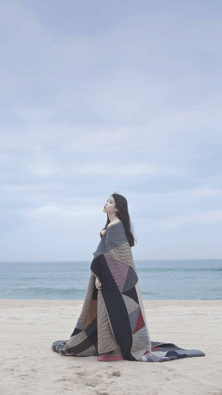drama 桌布 壁紙 — 一個女人裹著幾何圖案被子，平靜地坐在沙灘上，仰望柔和的多雲天空。