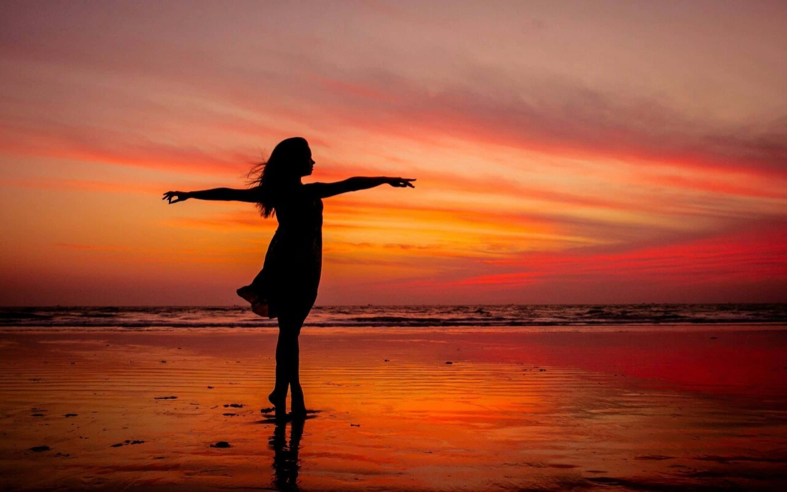 日落 桌布 壁紙 — 一個女性的剪影站在濕漉漉的海灘上，雙臂伸展，火熱的橙色和粉紅色日落色調塗滿天空並倒映在水面上。