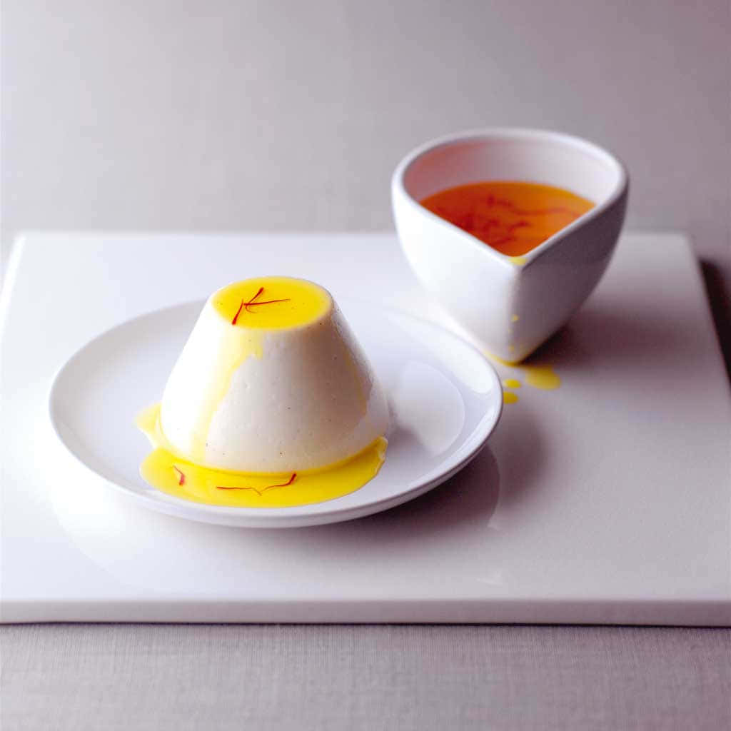 Blancmange 桌布 壁紙 — 一個奶油白色意式奶凍圓頂，上面綴有番紅花絲和金黃色醬汁，放在簡約的白色盤子上。