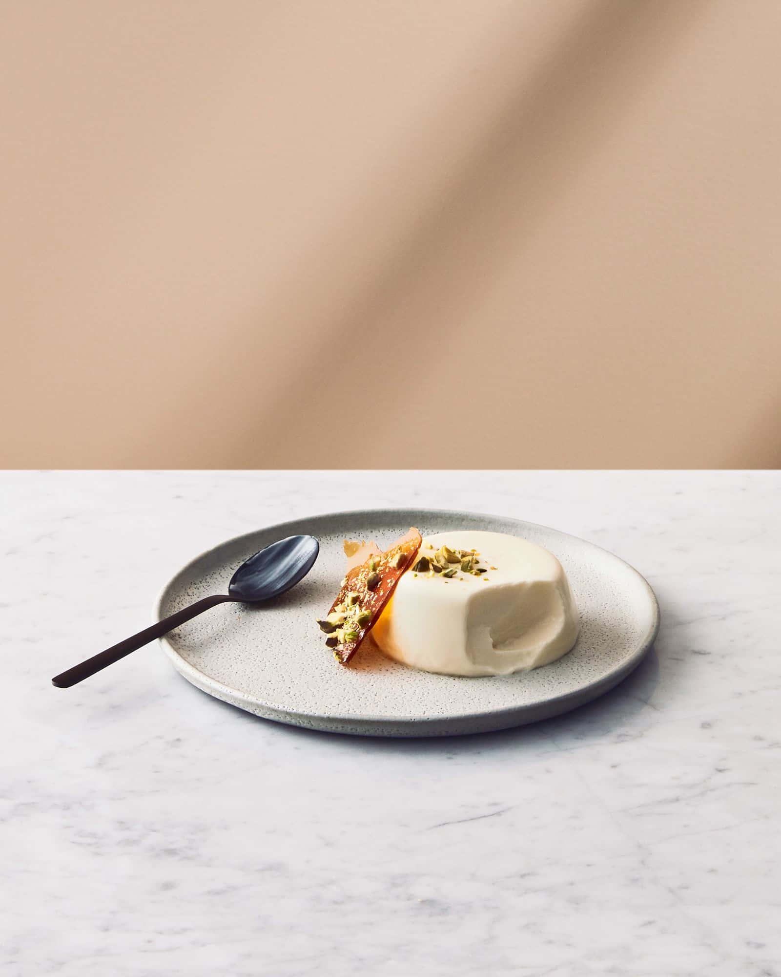 Minimalist Food 桌布 壁紙 — 一個奶油白色的意式奶凍，上面放有開心果和焦糖橙片，放在一個有斑點的盤子上，配上一把霧面黑湯匙，置於大理石表面上。