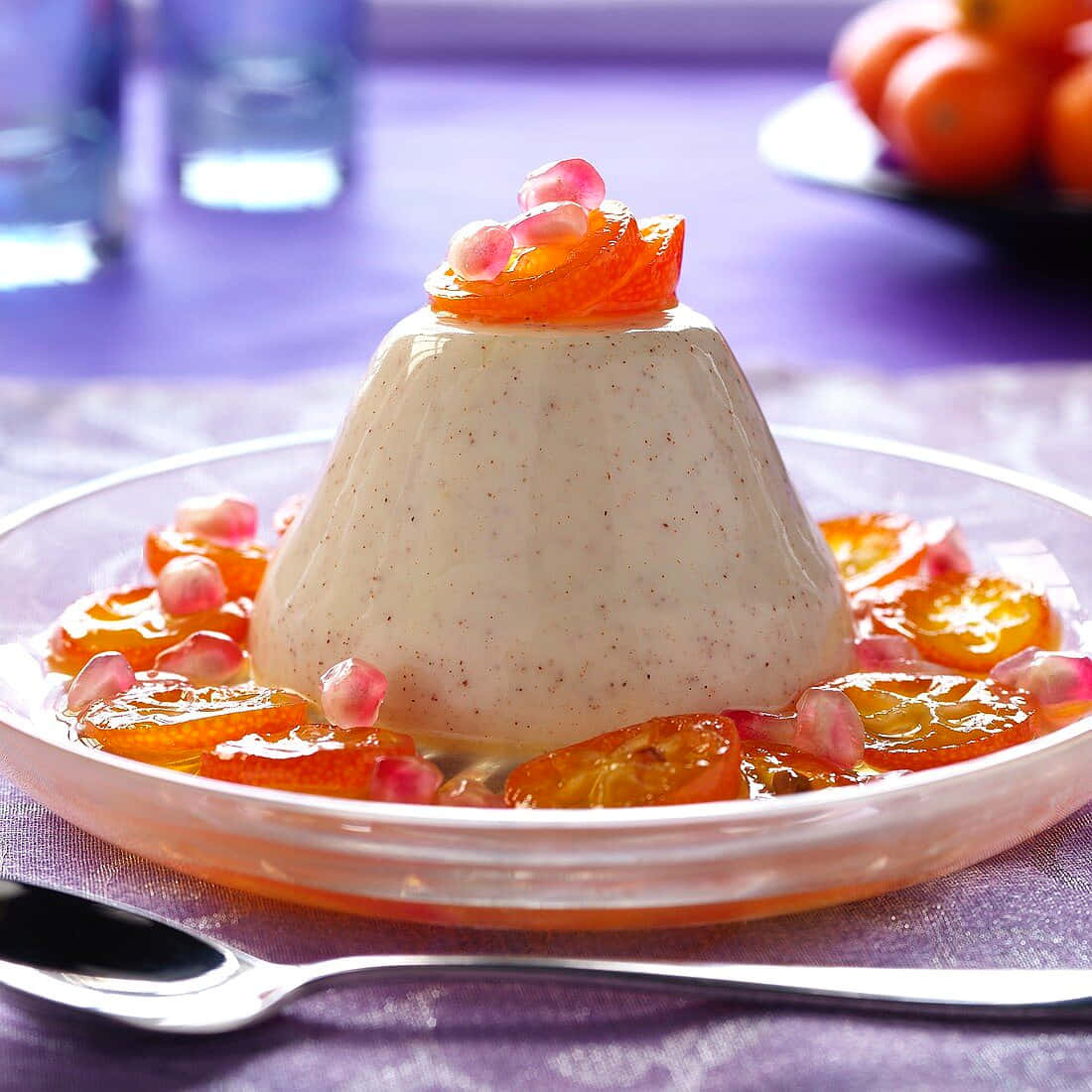 Blancmange 桌布 壁紙 — 一個奶油白色的意式奶凍圓頂，飾以蜜餞橙片和石榴籽，優雅地放在玻璃盤上，背景是柔和的紫色。