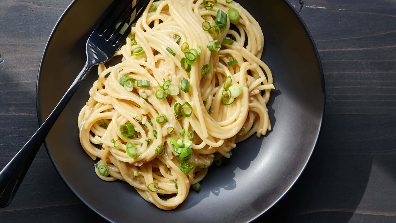Food Photography 桌布 壁紙 — 一個奶油義大利麵菜餚，上面撒有切碎的青蔥，放在一個深色碗裡，旁邊有一把叉子，置於木質表面上。