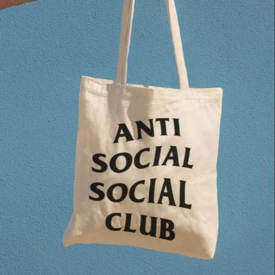 club 桌布 壁紙 — 一個奶油色帆布手提袋，上面有粗體黑色 'ANTI SOCIAL SOCIAL CLUB' 文字，掛在質感藍色牆上，在明亮日光下。