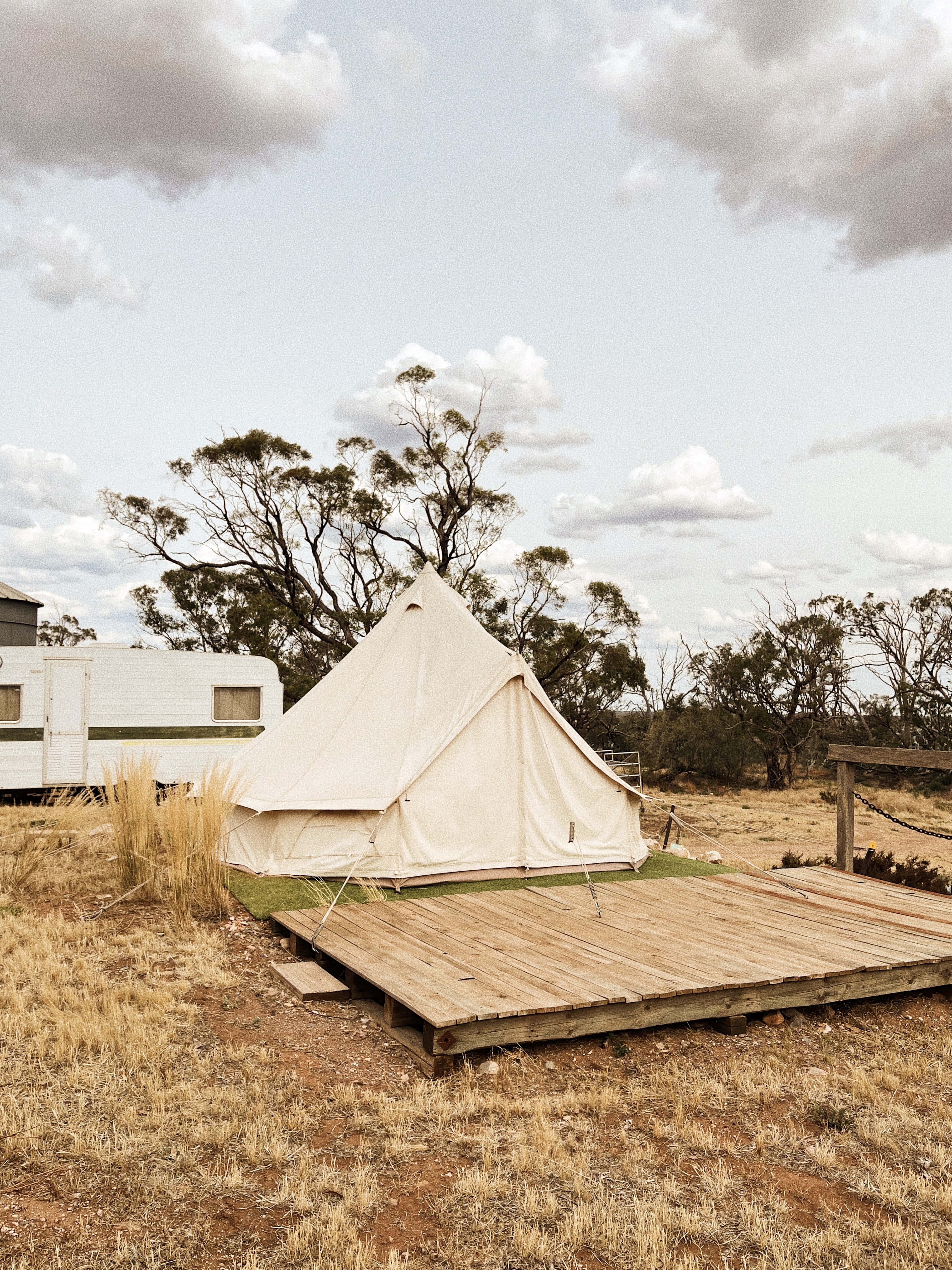 Camping 桌布 壁紙 — 一個奶油色鐘形帳篷放在木製甲板上，位於乾燥的草地，天空多雲，附近有散落的樹木和一輛露營車。