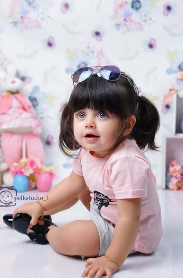 Baby Girl 桌布 壁紙 — 一個好奇的幼兒，梳著深色辮子，太陽眼鏡掛在頭上，坐在白色地板上，周圍是粉紅色花卉裝飾。
