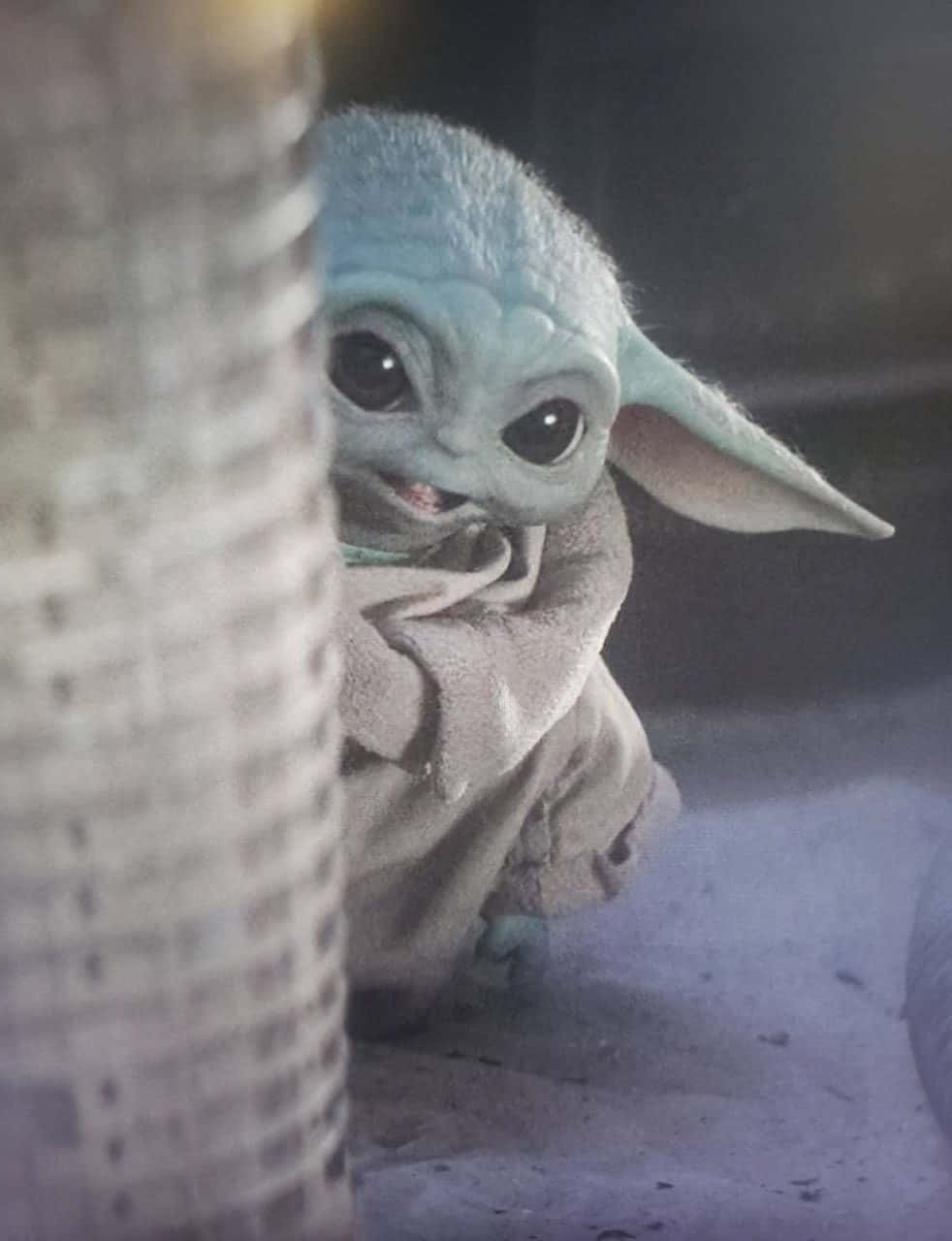 Baby Yoda Phone 桌布 壁紙 — 一個好奇的綠色皮膚嬰兒尤達在昏暗燈光下從編織柱子後面探出頭來。