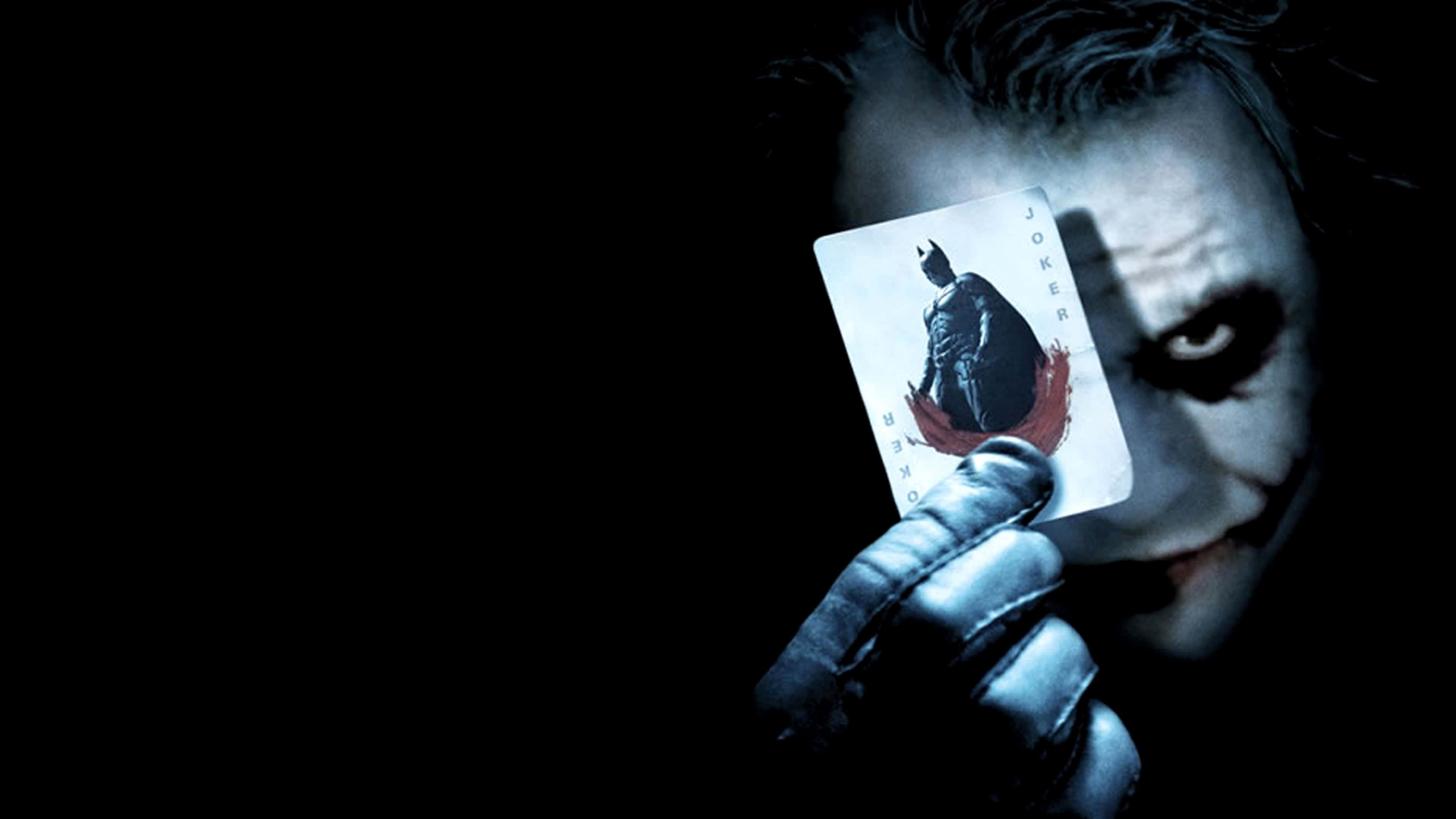 Dark Knight Joker In 4K Ultra HD 桌布 壁紙 — 一個威脅性的小丑拿著一張帶有蝙蝠俠剪影的小丑牌，背景陰暗。
