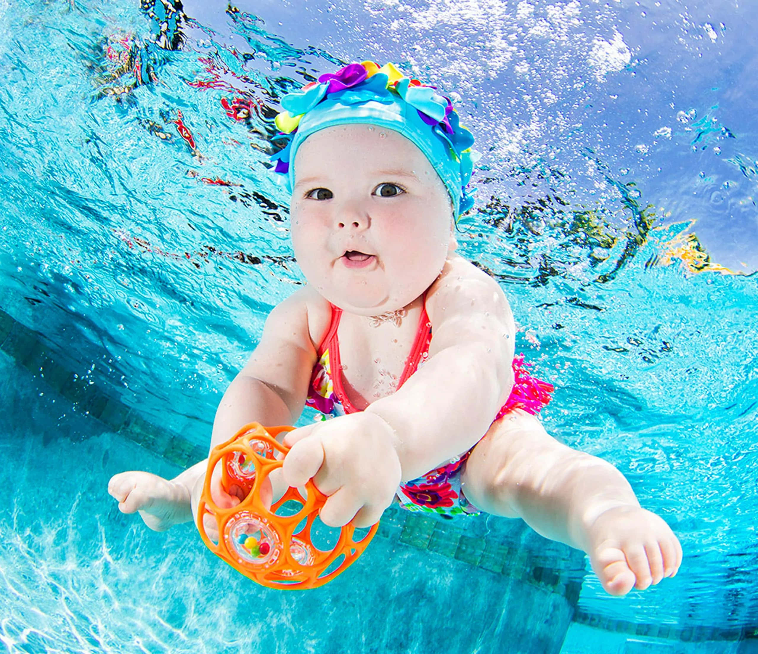 Swimming 桌布 壁紙 — 一個嬰兒在泳池水底，戴著花卉圖案泳帽，手持橙色玩具，向上看。