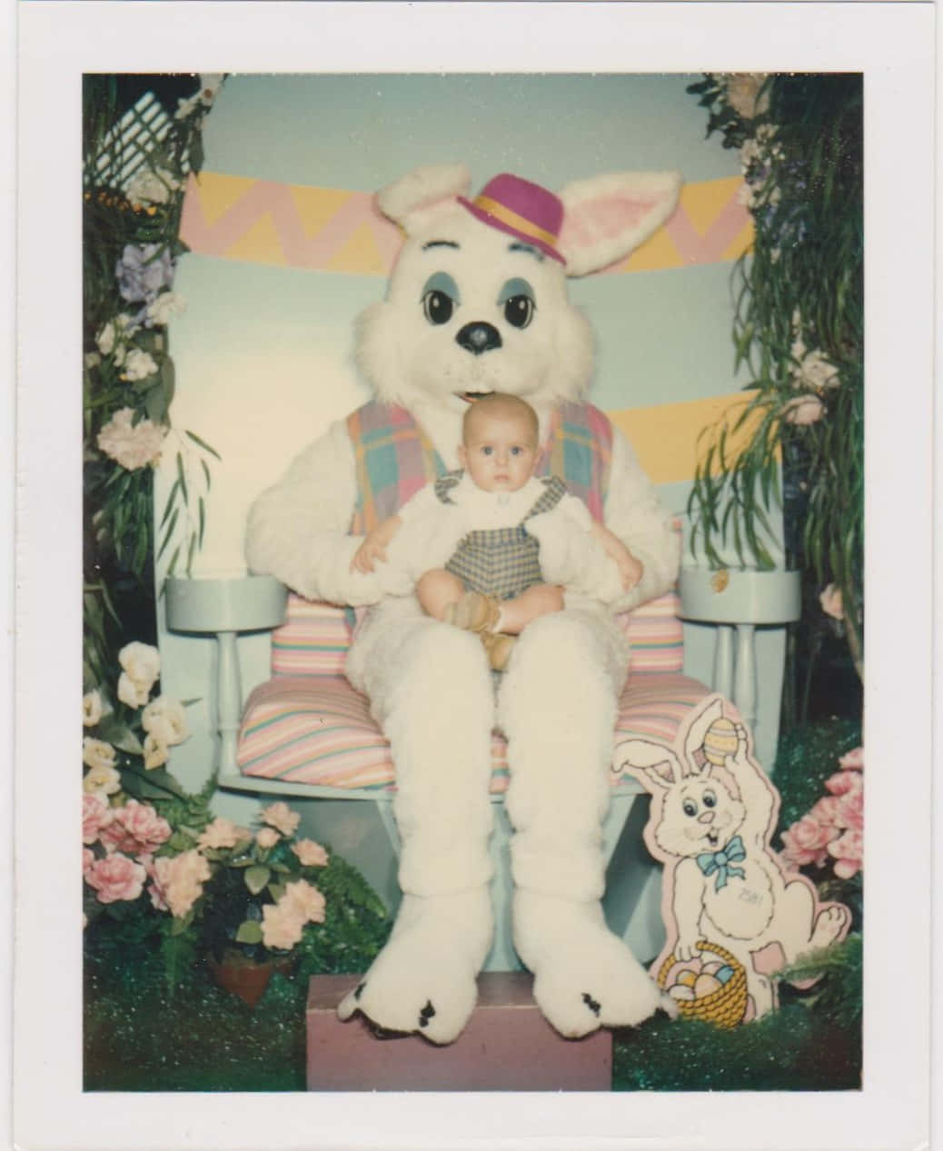 Creepy Easter Bunny 桌布 壁紙 — 一個嬰兒坐在一個大型白色復活節兔子上，位於一個復古照相亭內，周圍環繞著粉紅色的花朵和節日裝飾。