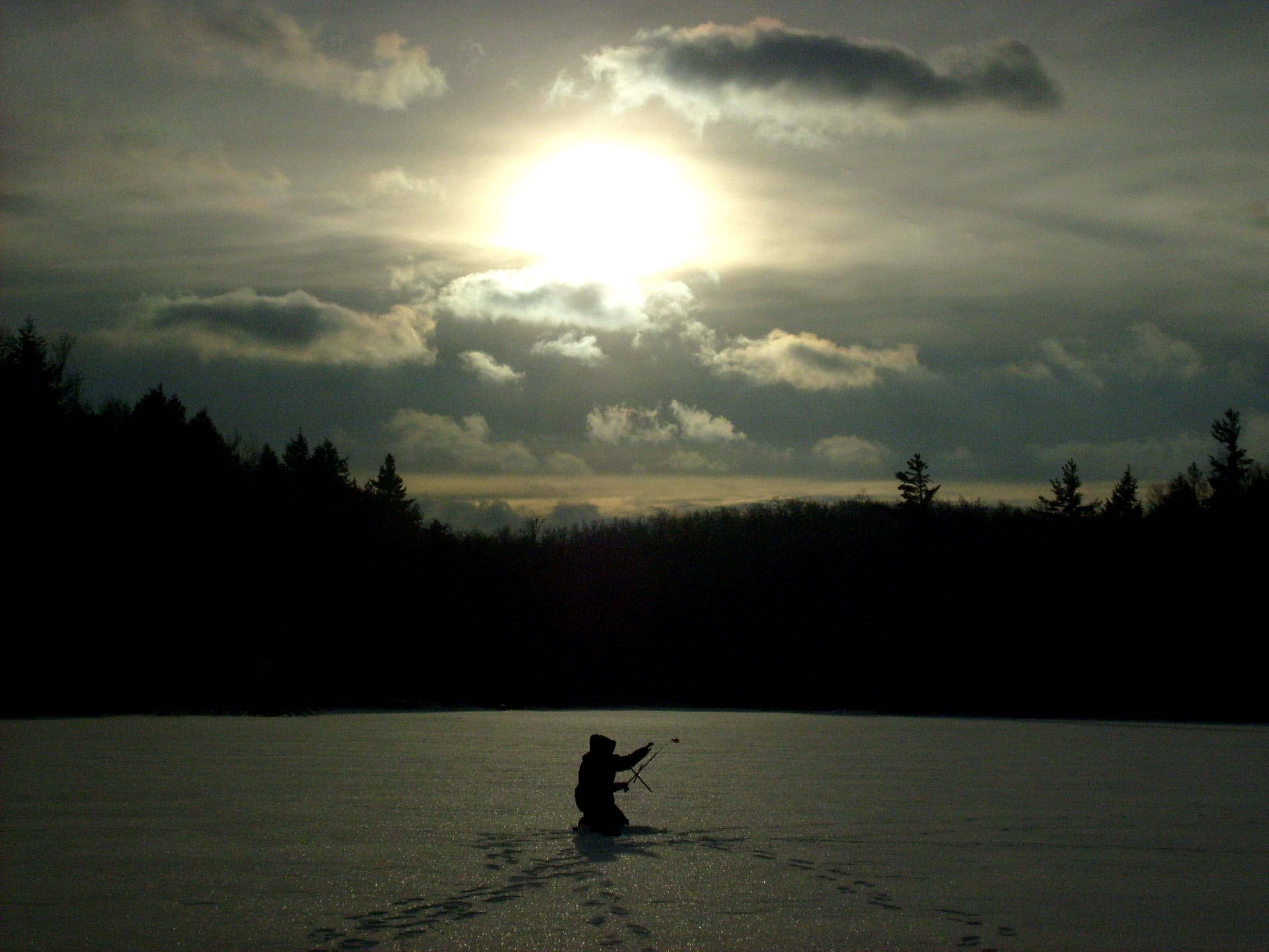 Ice Fishing 桌布 壁紙 — 一個孤獨漁夫的剪影跪在冰凍的湖面上，明亮的陽光穿透厚重的雲層。