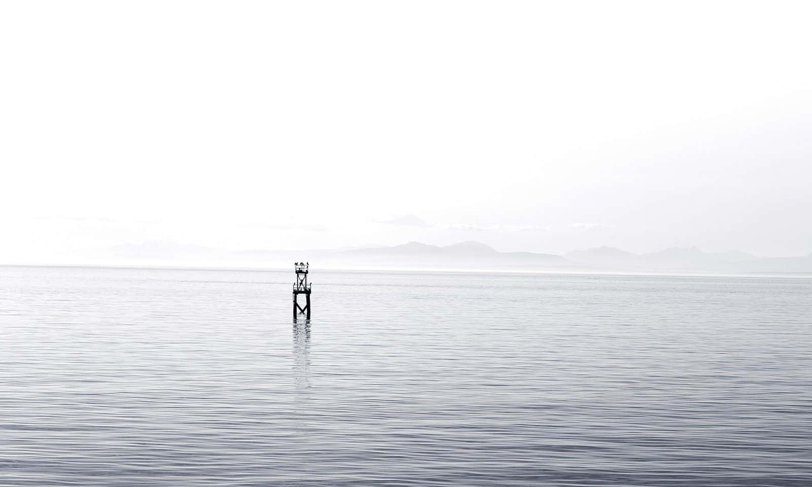 Black And White Ocean 桌布 壁紙 — 一個孤獨的、風化的浮標矗立在平靜的灰色水面上，天空多雲，遠處有朦朧的山脈。