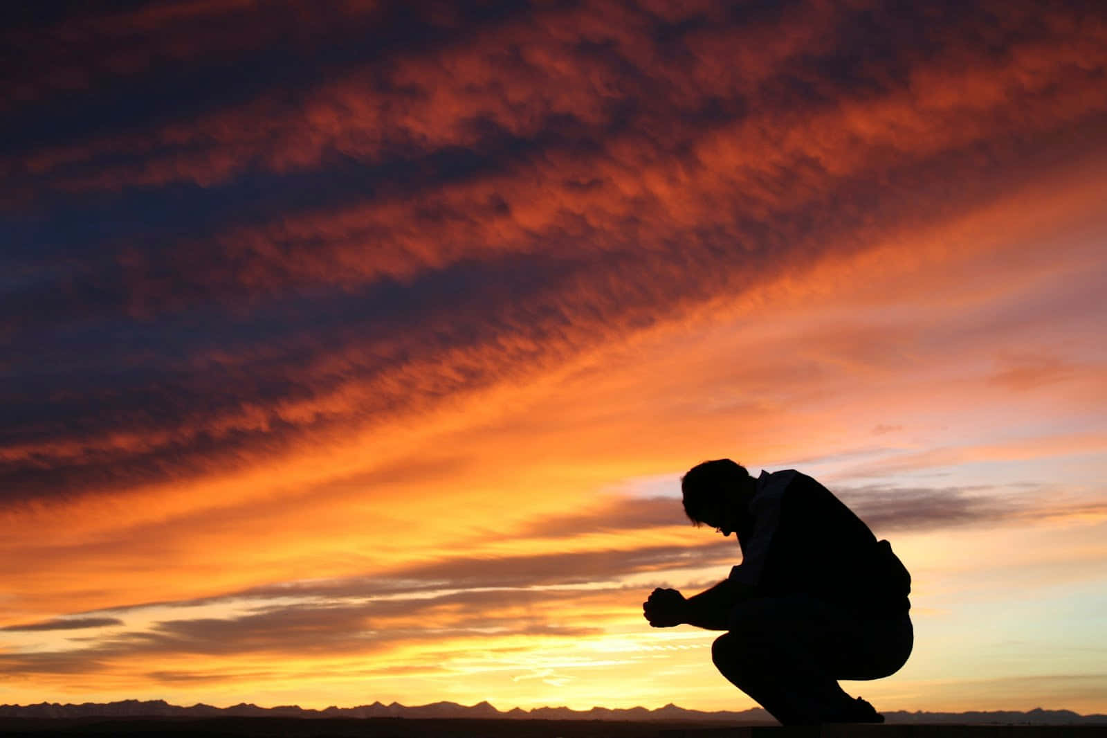 Praying 桌布 壁紙 — 一個孤獨的人以剪影跪在熾熱的日落天空前，天空有戲劇性的橙色和紫色雲彩。