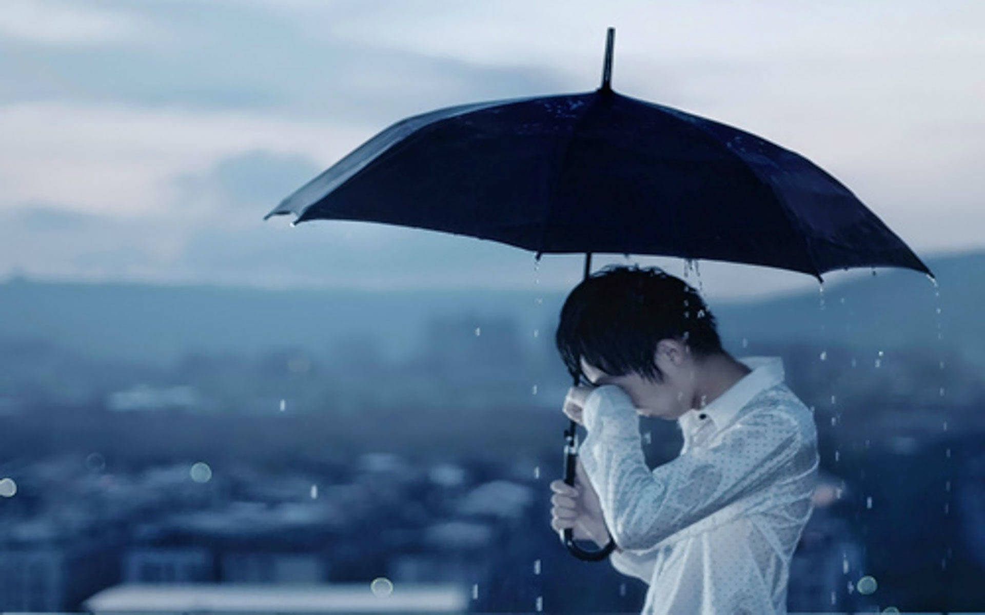Sad Alone 桌布 壁紙 — 一個孤獨的人在一把黑色雨傘下避雨，雨落在黃昏時模糊的城市景觀上。