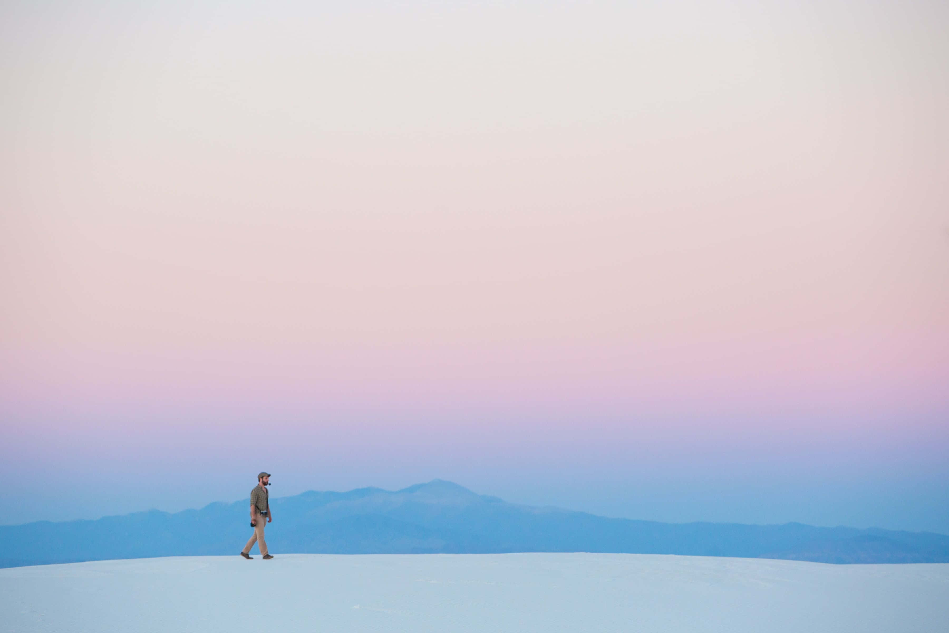 Minimalist Ocean 桌布 壁紙 — 一個孤獨的人在一片廣闊的白色沙漠中行走，天空是柔和的粉紅色和藍色暮色，遠處有山脈。