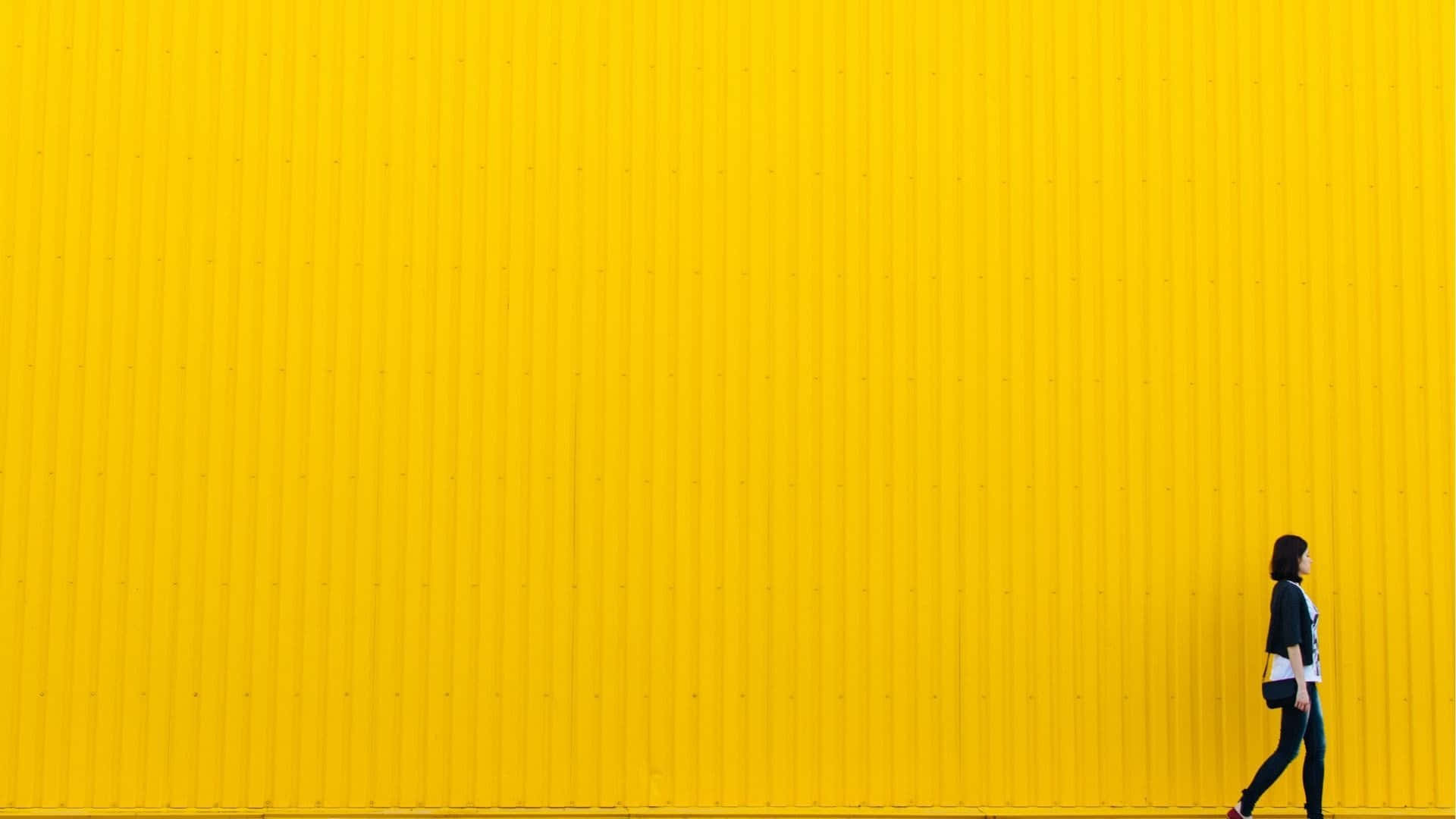 Canva 桌布 壁紙 — 一個孤獨的人在明亮的日光下走過一座高聳、鮮黃色的波紋牆。