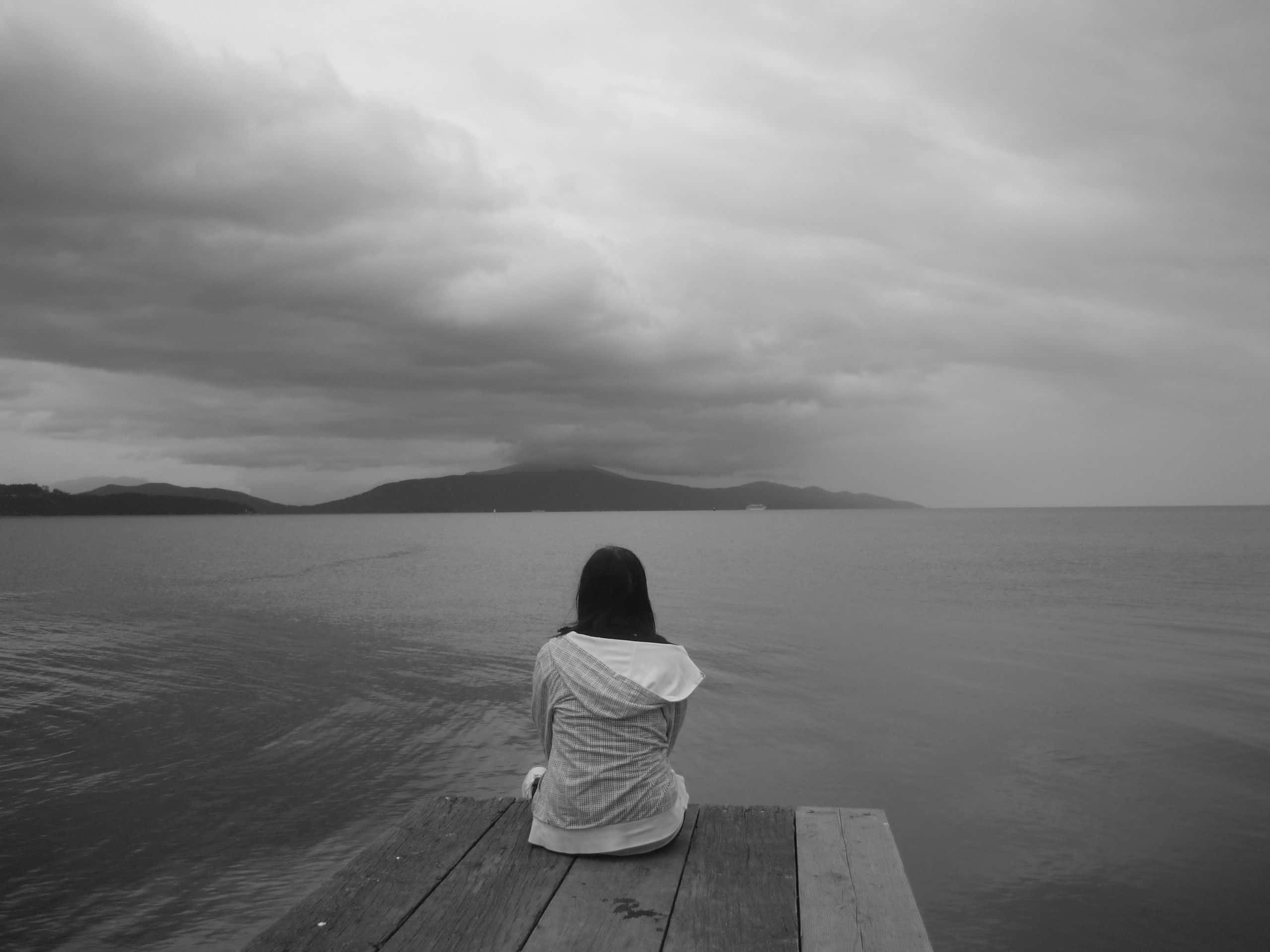 Alone 桌布 壁紙 — 一個孤獨的人坐在木碼頭上，凝視著陰天下一片廣闊平靜的水面，遠方有山巒。