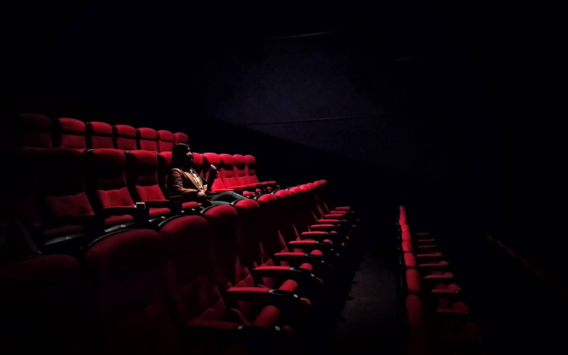 Alone 桌布 壁紙 — 一個孤獨的人坐在漆黑的電影院裡，周圍是一排排空著的紅色座位，在昏暗的環境光下。