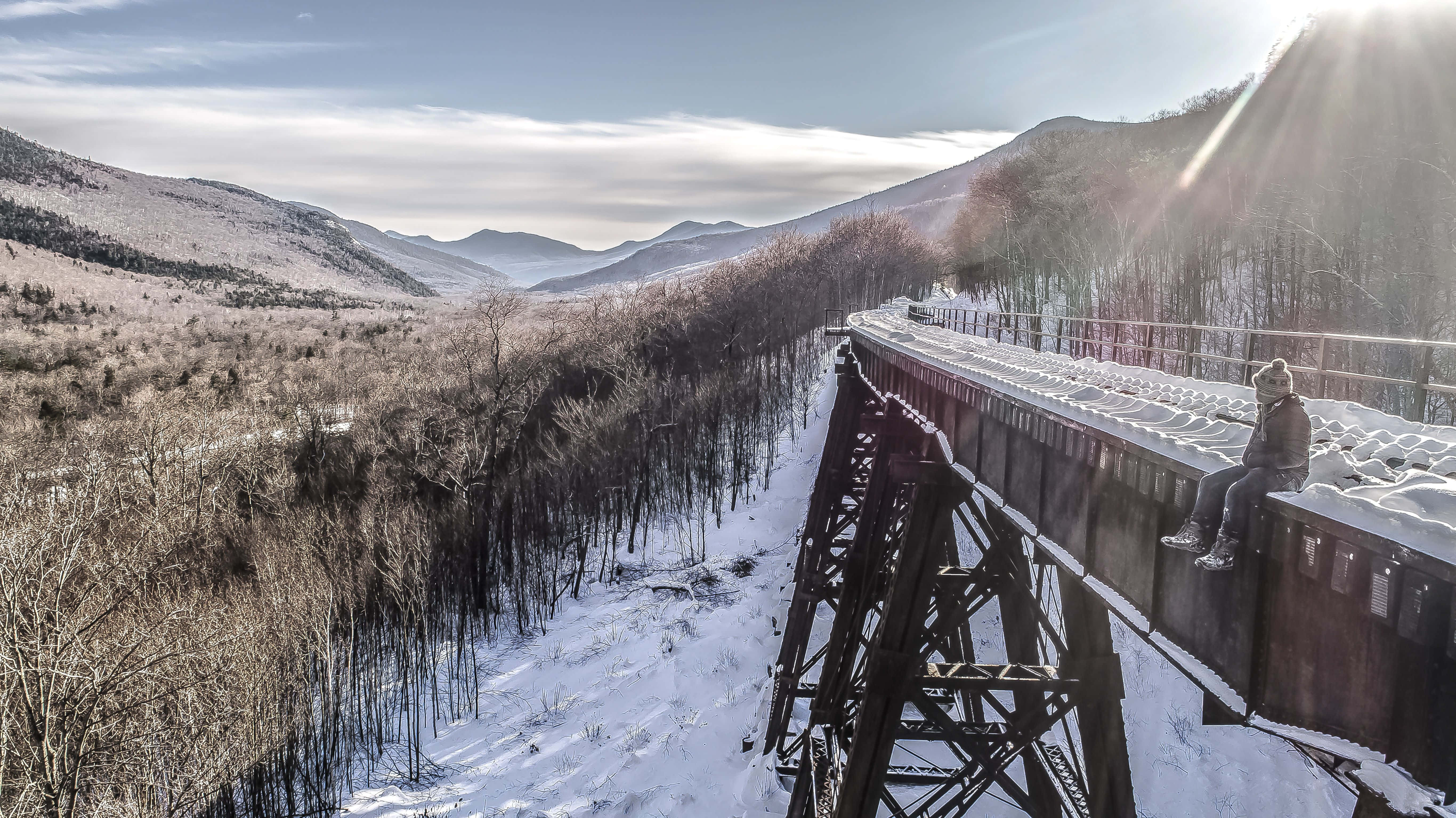 Winter Landscape 桌布 壁紙 — 一個孤獨的人坐在被雪覆蓋的鐵路橋上，俯瞰著廣闊的冬季山谷，陽光穿過雲層照射下來。