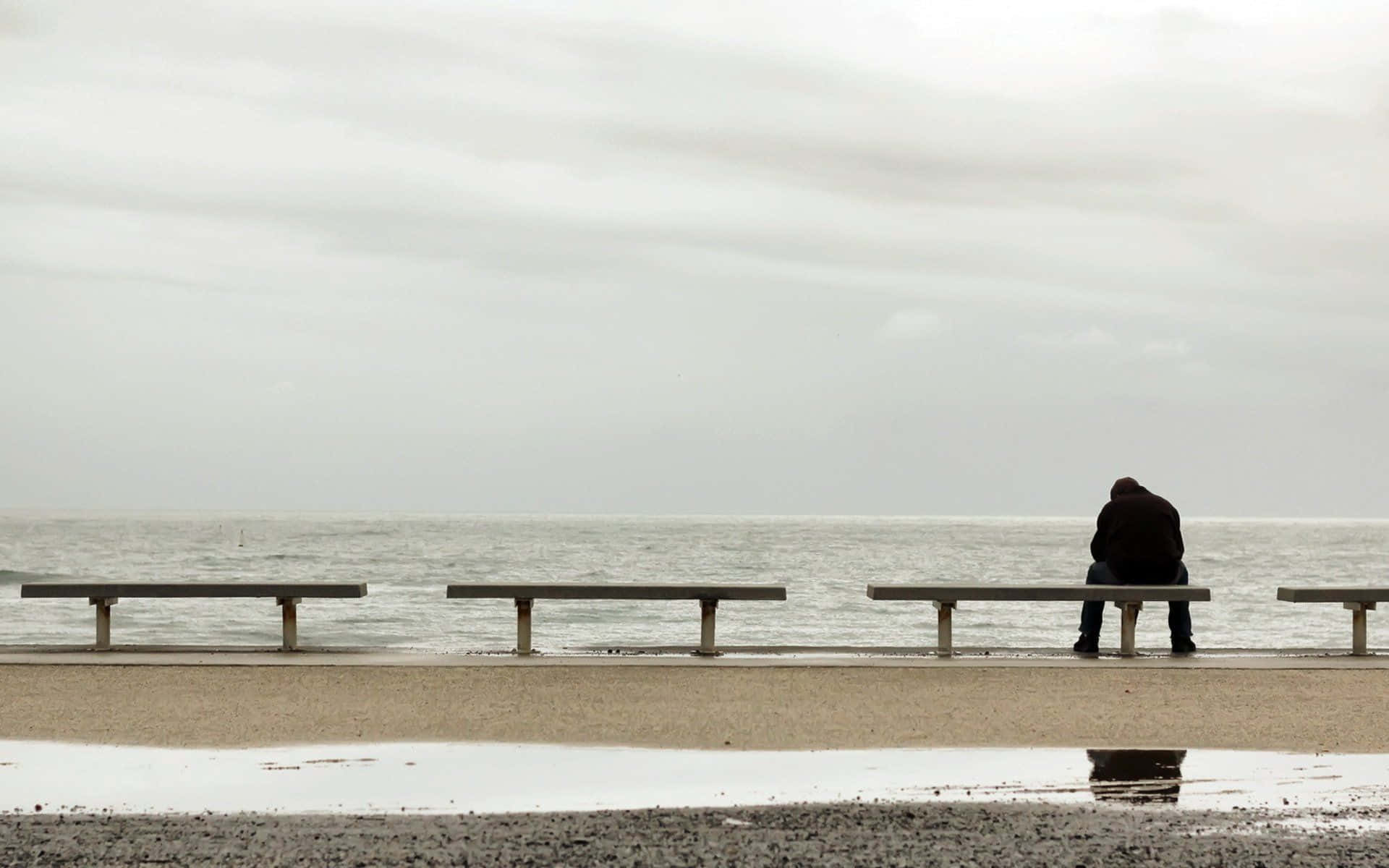 Alone 桌布 壁紙 — 一個孤獨的人彎腰坐在長椅上，俯瞰著陰天下一片廣闊的灰色海洋。