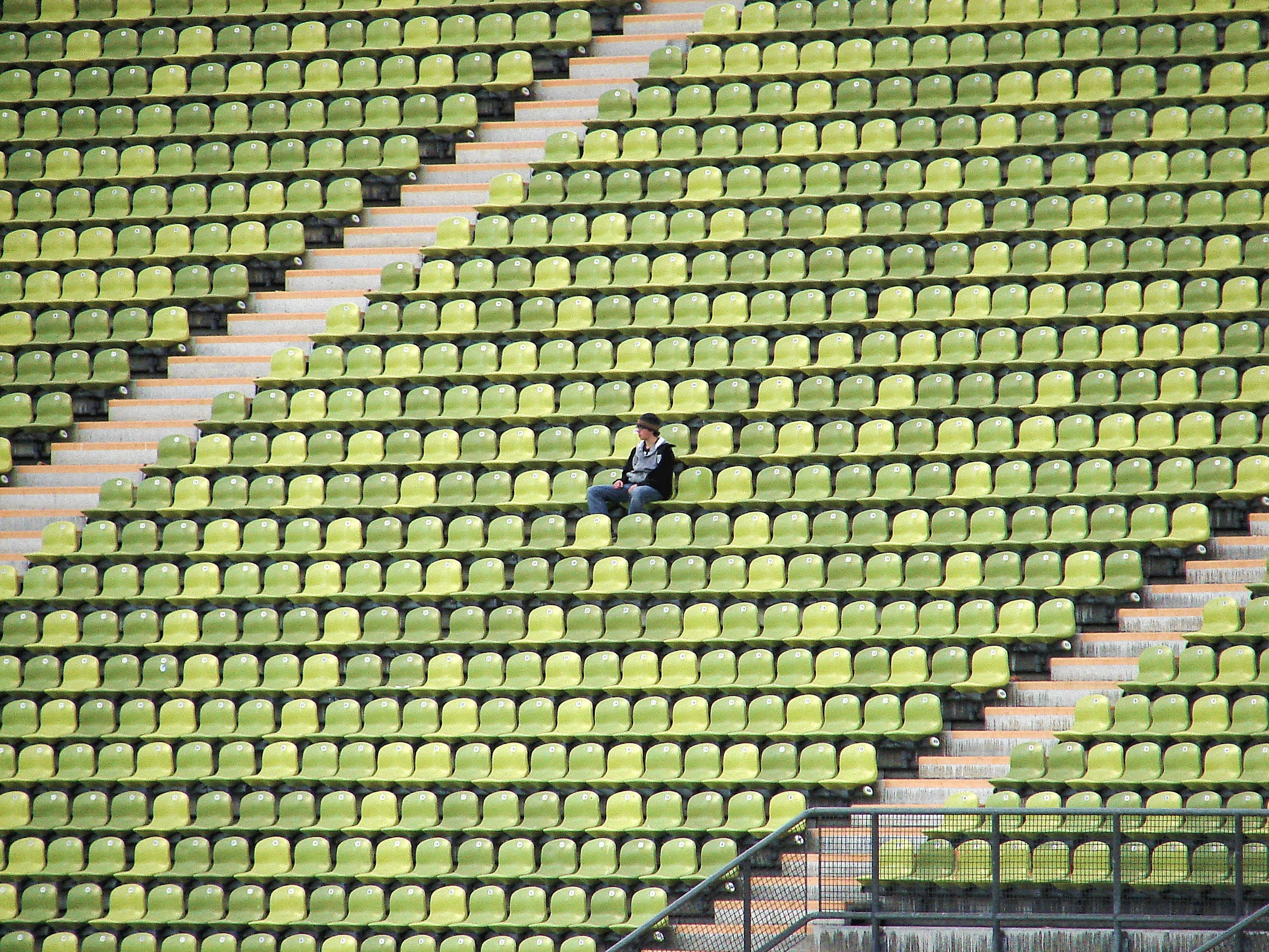 Alone 桌布 壁紙 — 一個孤獨的人獨自坐在綠色體育場座位上，周圍是空蕩蕩的排位，在日光下。