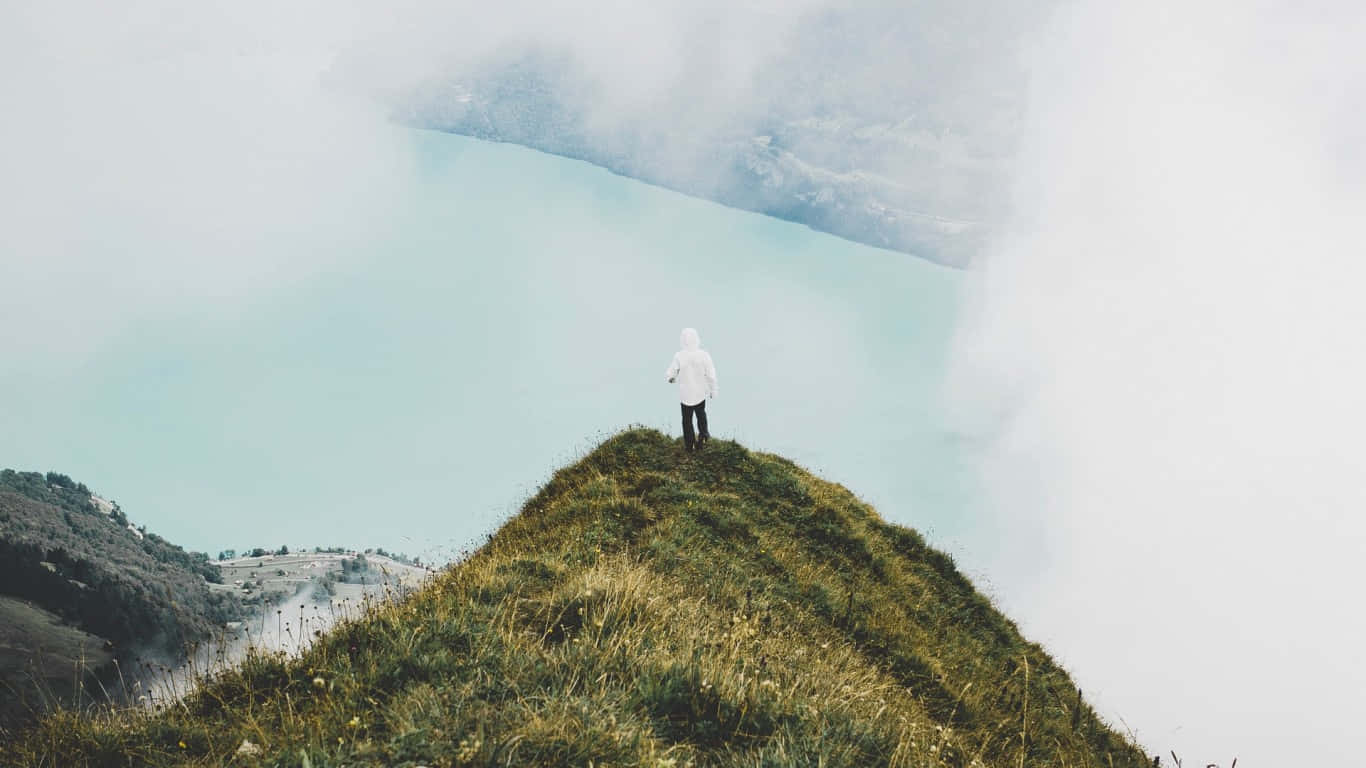 Traveler 桌布 壁紙 — 一個孤獨的人穿著白色連帽衫，站在長滿草的山脊上，俯瞰著霧氣繚繞的綠松石色湖泊。