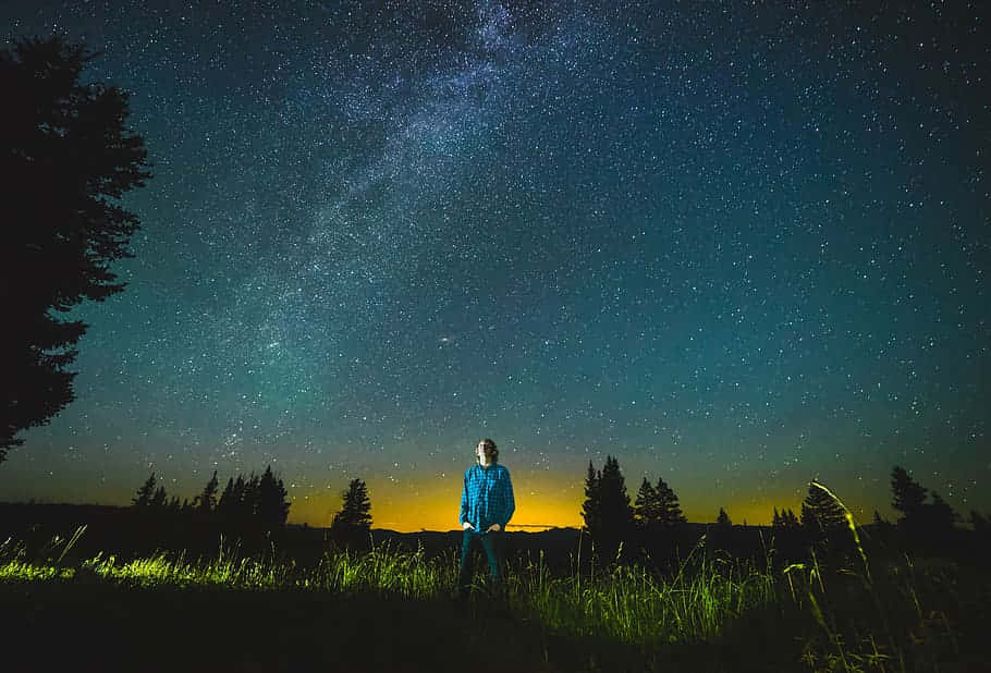 Stars 桌布 壁紙 — 一個孤獨的人穿著藍色外套，站在草地上，頭頂是令人屏息的星空，銀河在閃耀。