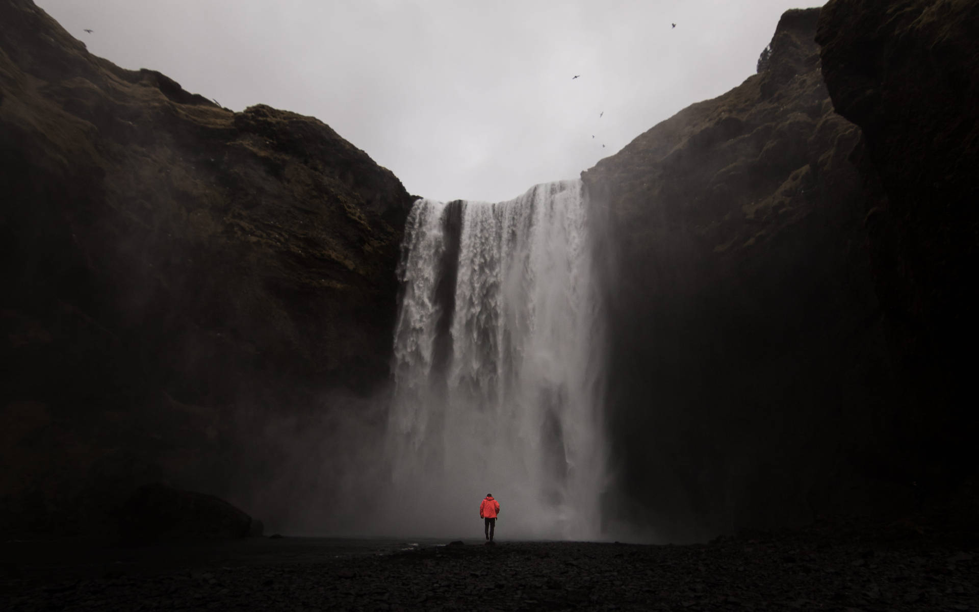 Alone 桌布 壁紙 — 一個孤獨的人穿著鮮橙色外套，站在陰天下黑暗懸崖之間傾瀉而下的巨大瀑布前。