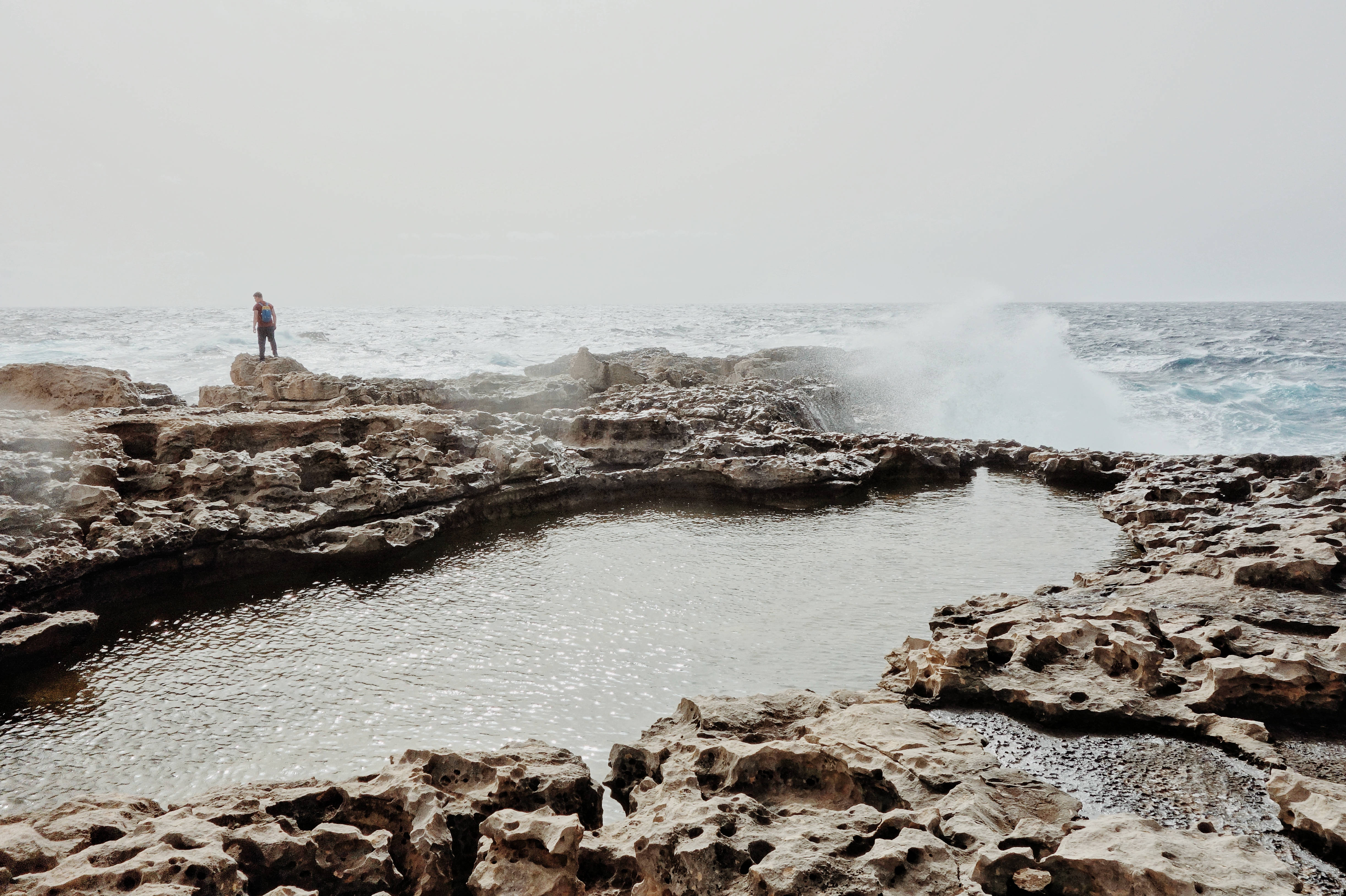 Malta 桌布 壁紙 — 一個孤獨的人站在崎嶇的海岸岩石上，海浪在陰天之下衝入天然岩石池。
