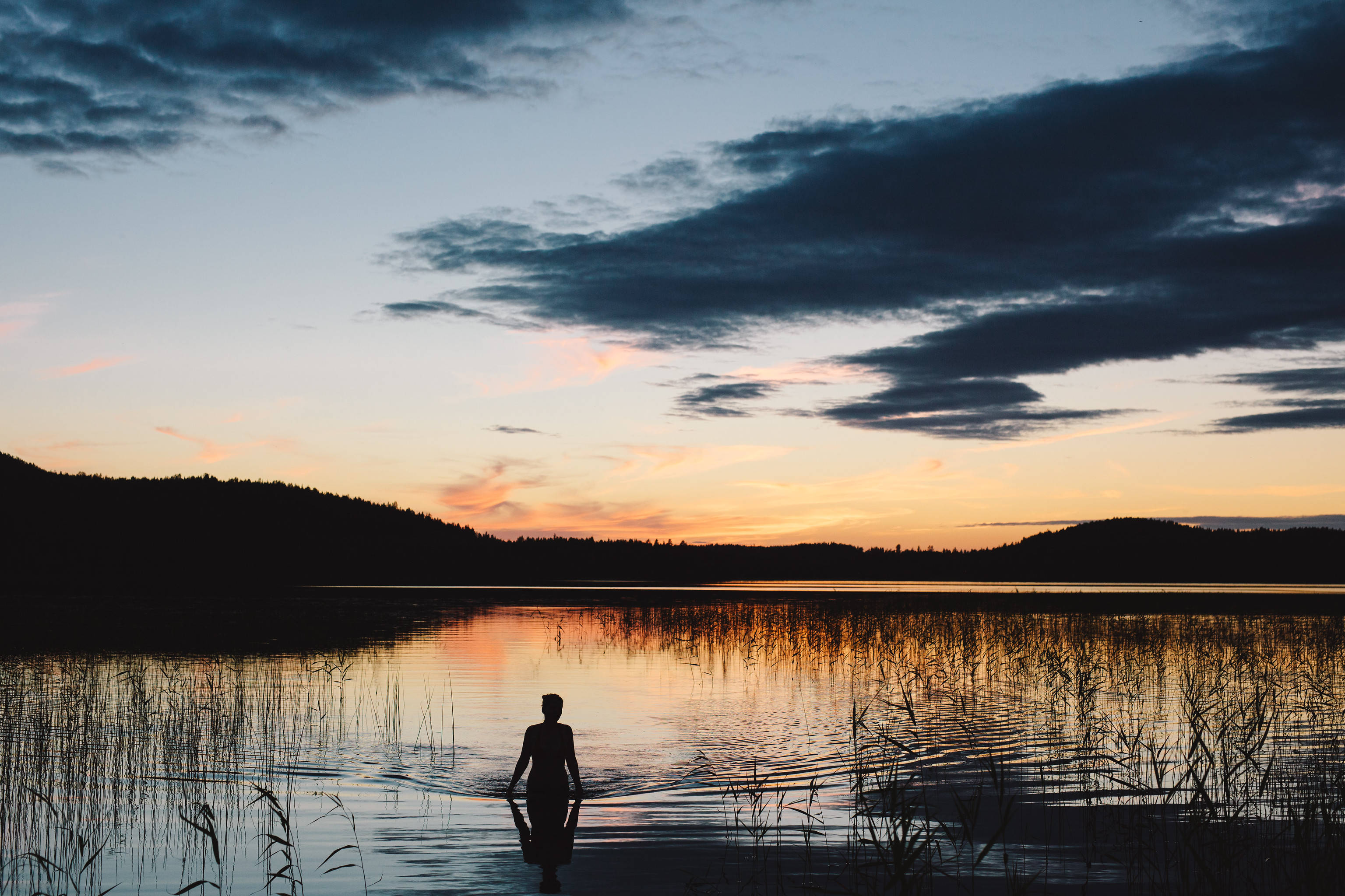 Sweden 桌布 壁紙 — 一個孤獨的人站在日落時分的淺水中，剪影映襯著橙色和深藍色雲朵的天空，倒映在平靜的湖面上。