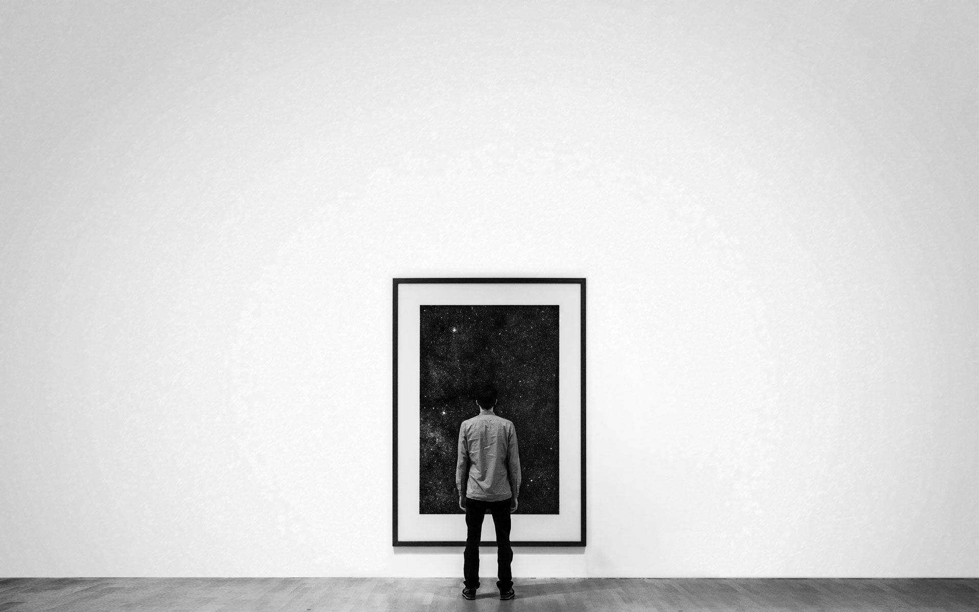 Gallery 桌布 壁紙 — 一個孤獨的人站在極簡主義畫廊中，凝視著純白牆上一個裱框的黑白星空印刷品。