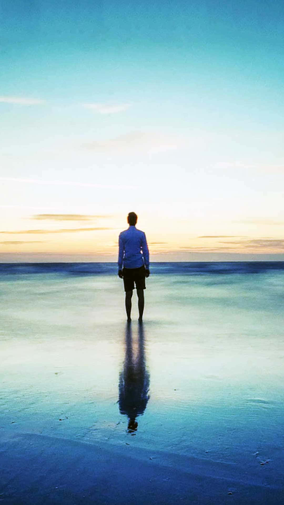 桌布 壁紙 — 一個孤獨的人站在水邊，夕陽西下，他們的剪影映照在平靜的海洋上，天空呈現漸變色彩。