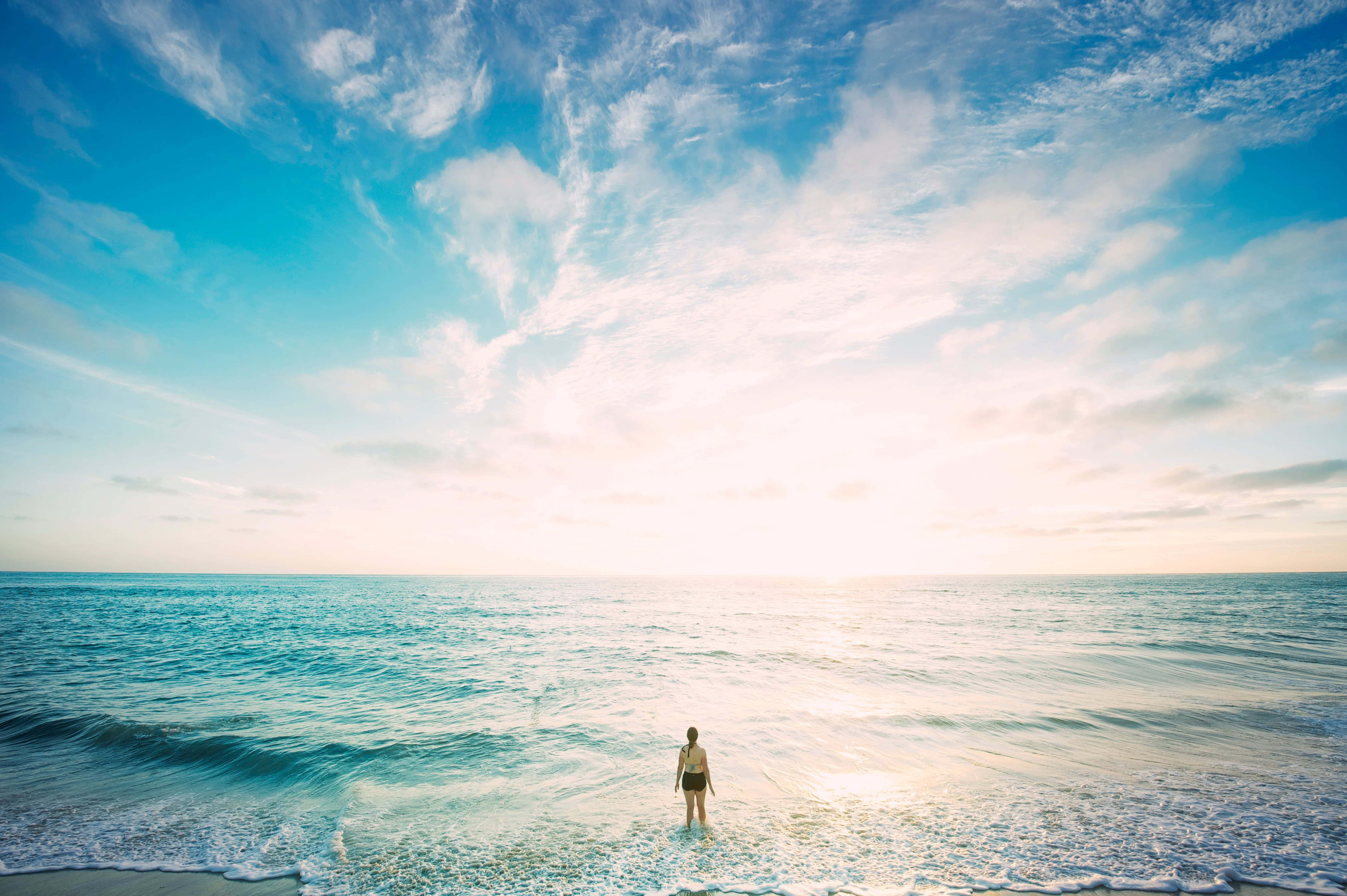 Beautiful Ocean 桌布 壁紙 — 一個孤獨的人站在海岸線上，溫柔的波浪輕拍他們的腳，廣闊的天空被柔和的雲彩和金黃的日落色調所渲染。