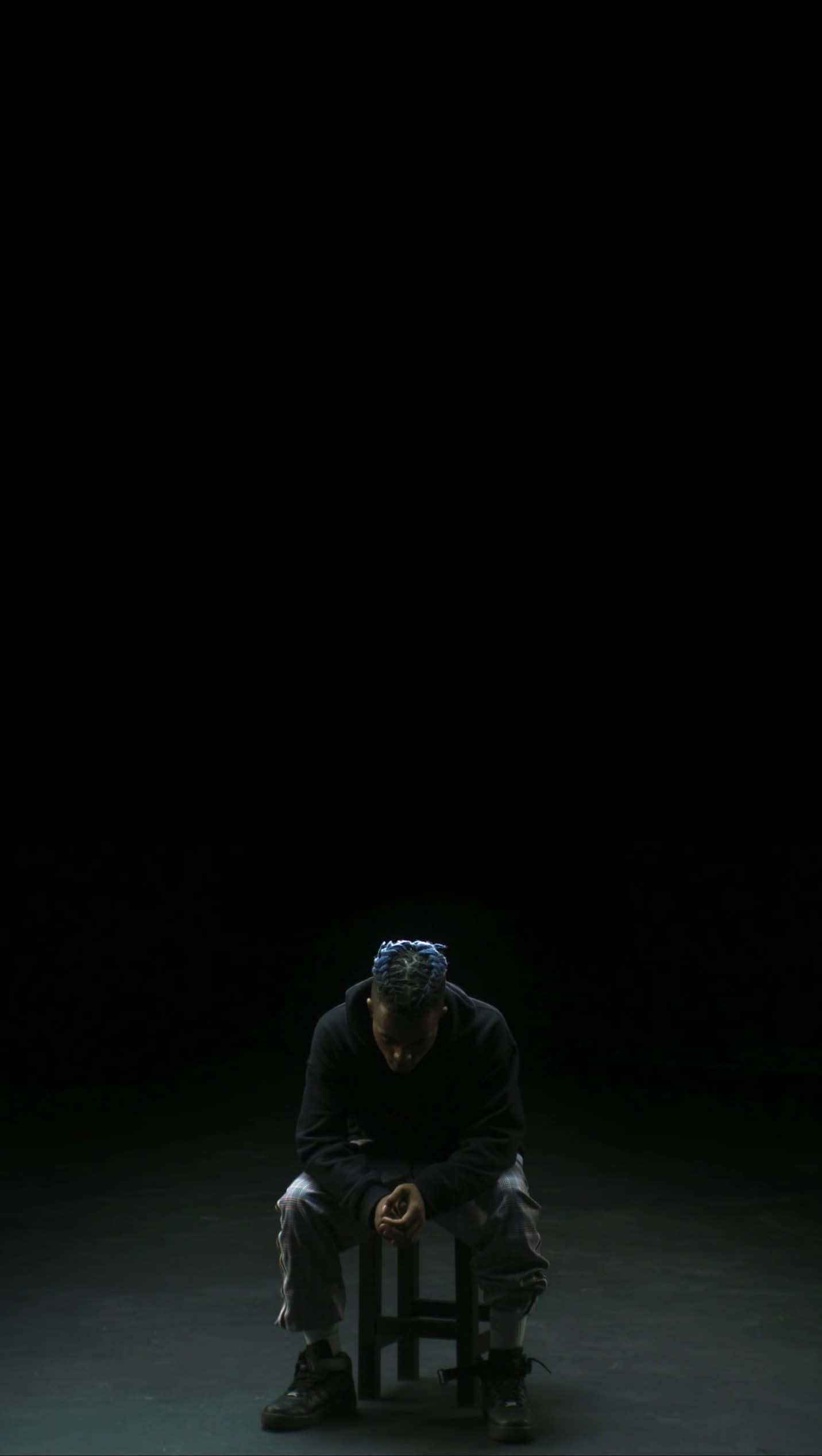 暗色美學 iPhone 桌布 壁紙 — 一個孤獨的人蜷縮在凳子上，身處漆黑虛空，被單一聚光燈照亮，投射出戲劇性的陰影。
