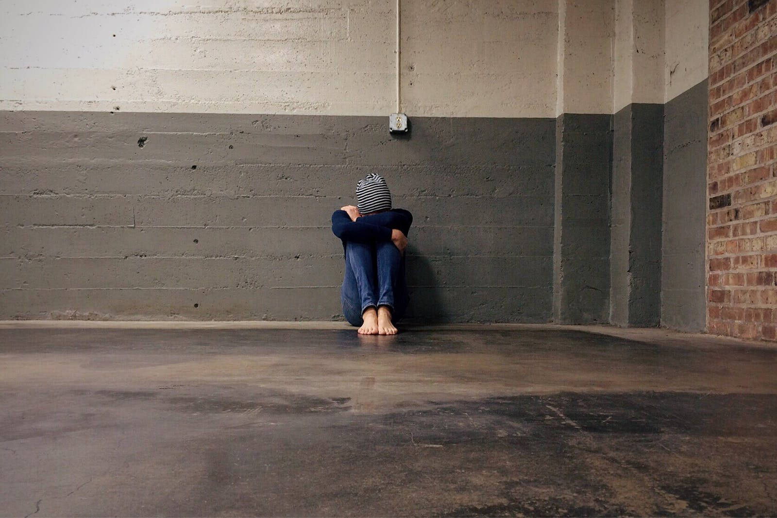 Lonely 桌布 壁紙 — 一個孤獨的人蜷縮在空蕩蕩的工業空間的混凝土牆邊，頭埋在膝蓋裡，戴著條紋毛帽和牛仔褲。