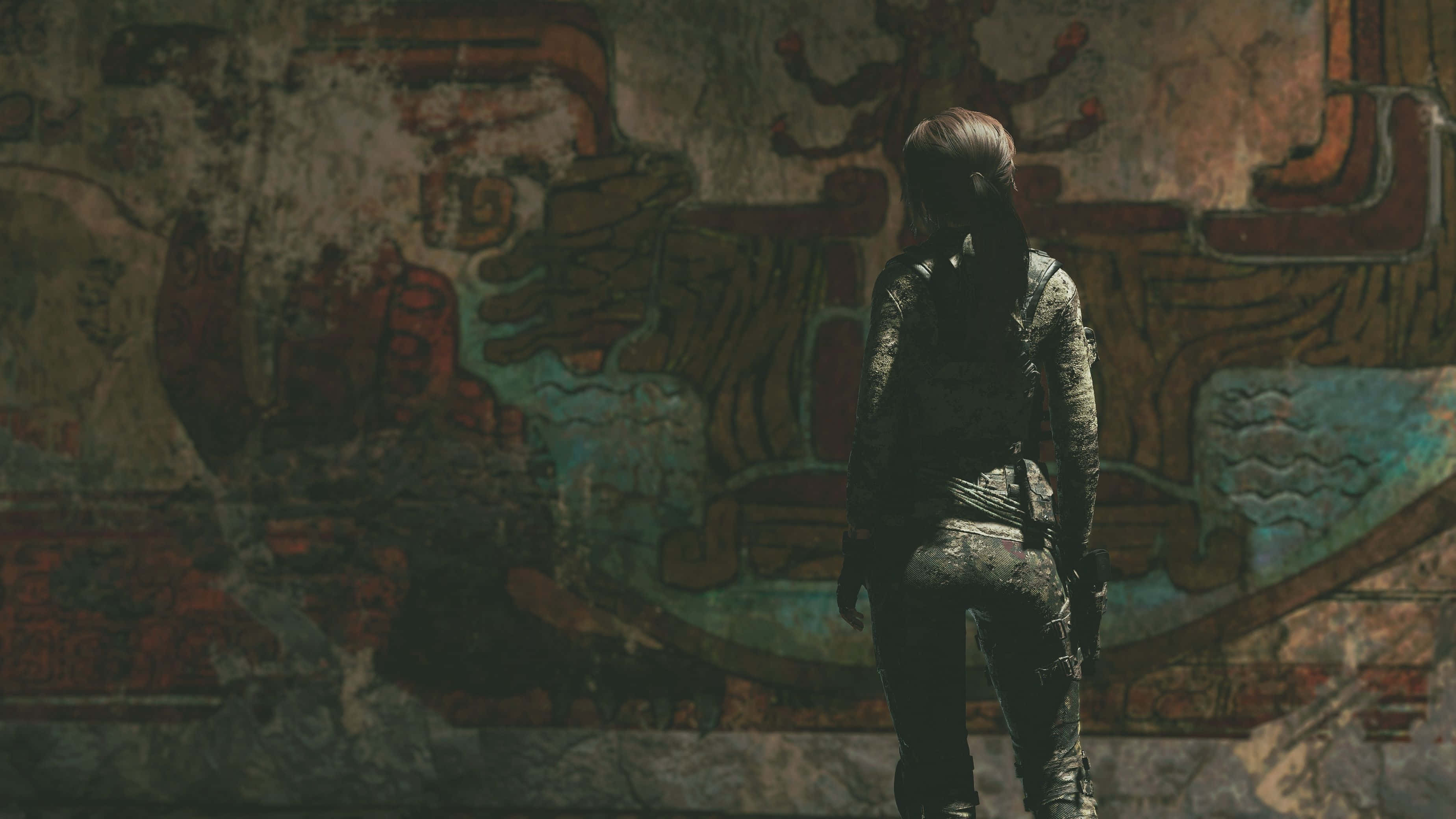 Lara Croft 桌布 壁紙 — 一個孤獨的冒險家穿著破舊裝備，站在一個昏暗廢墟中褪色的古代壁畫前，壁畫上有紅色和藍綠色的符文。