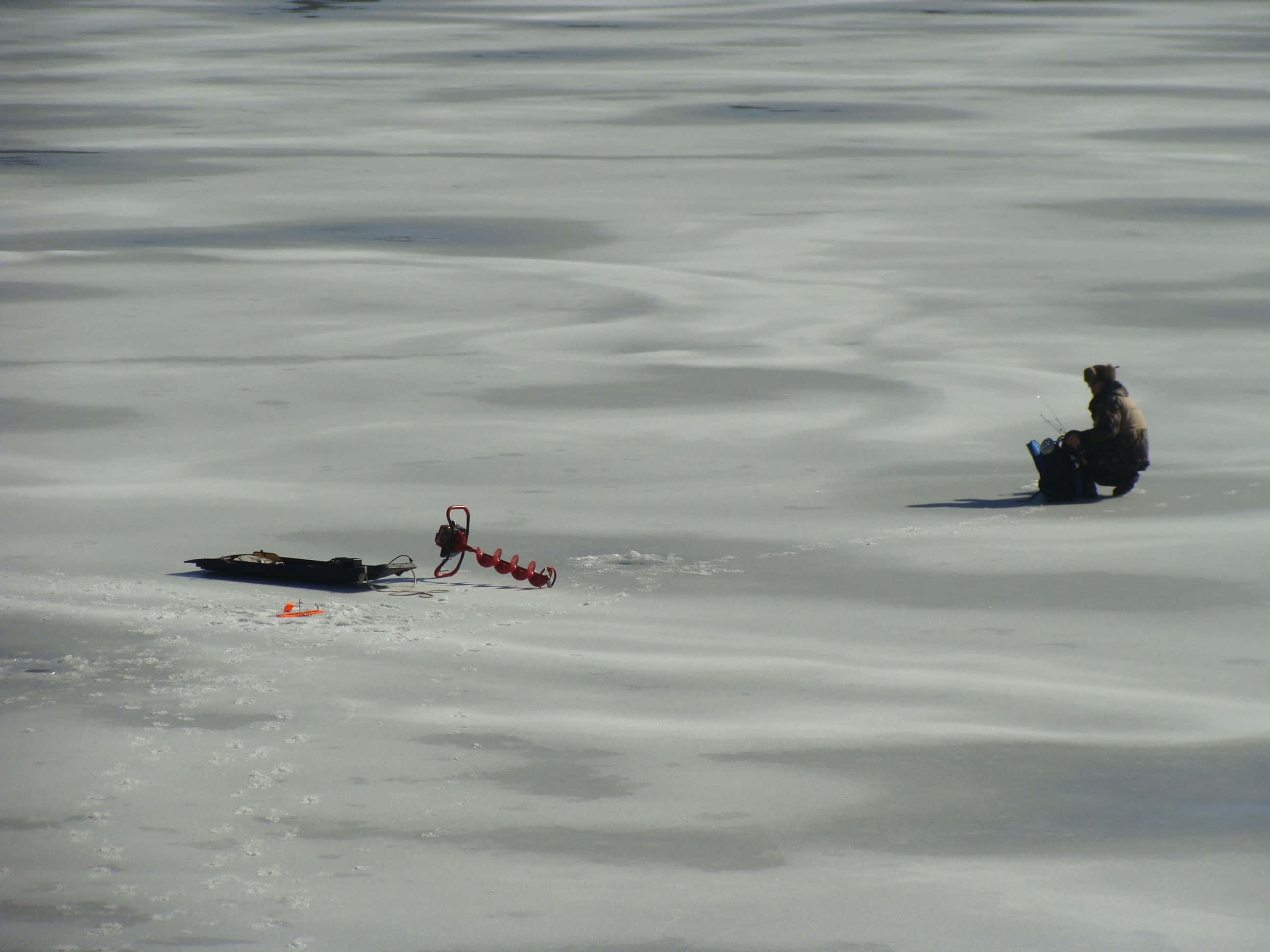 Ice Fishing 桌布 壁紙 — 一個孤獨的冰釣者坐在結冰的湖面上，附近有一個紅色冰鑽，在明亮的日光下。