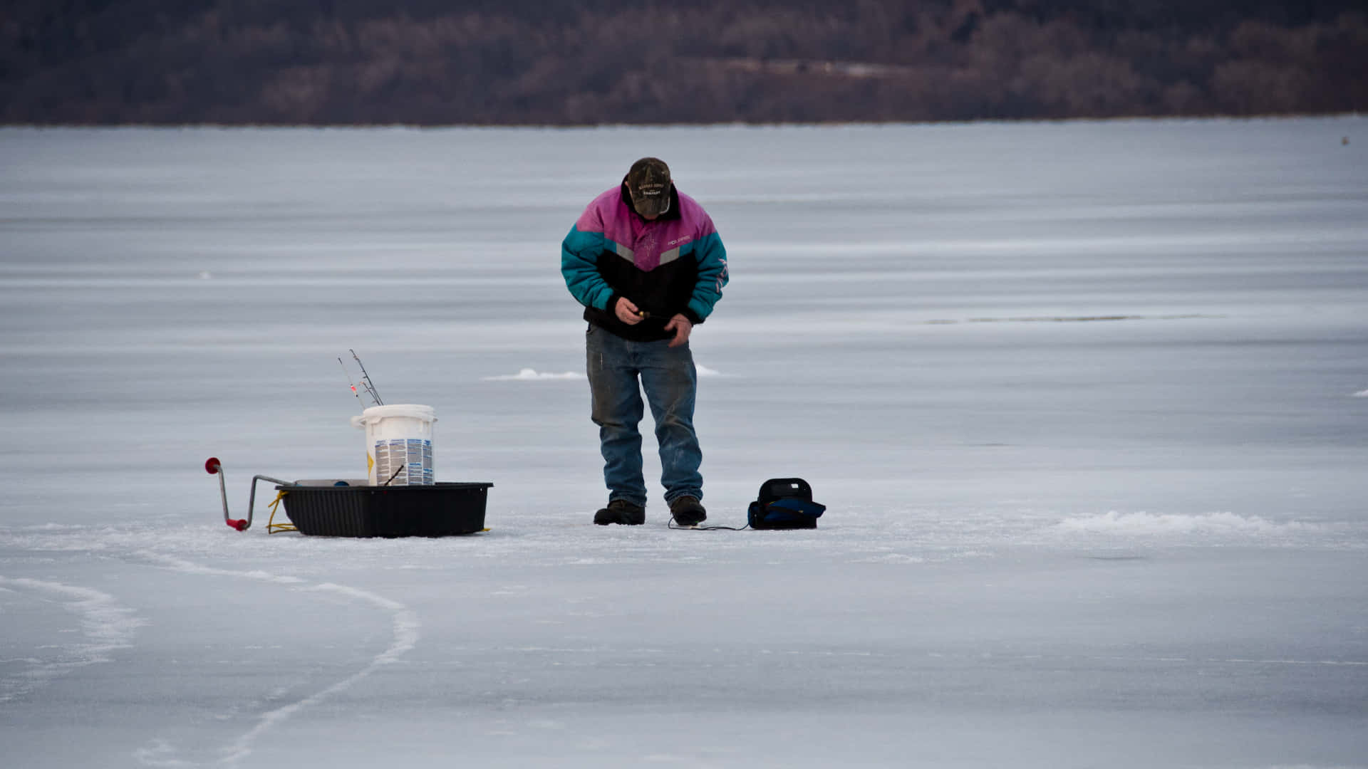 Ice Fishing 桌布 壁紙 — 一個孤獨的冰釣者穿著色彩繽紛的夾克，站在結冰的湖面上，在一個黑色冰釣裝置旁邊調整他的裝備。