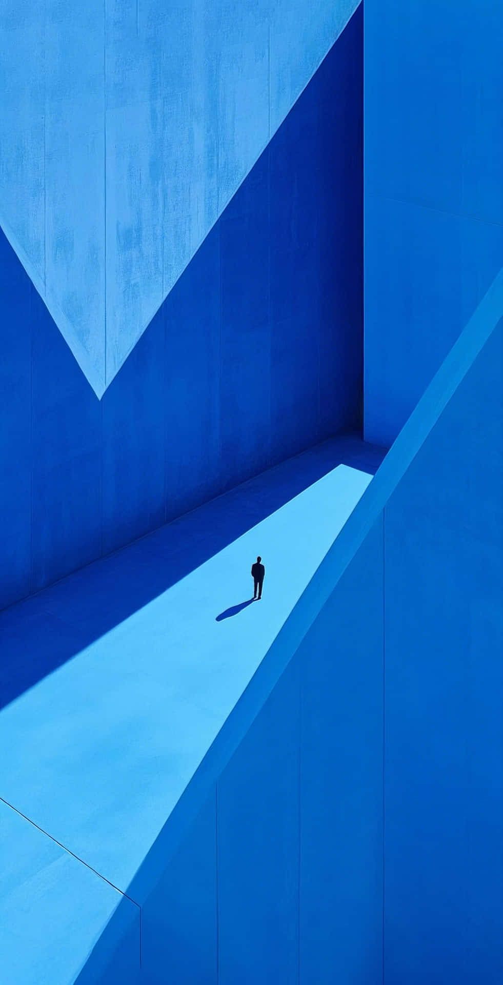 minimalist blue panels and solitary figure in sharp shadow 壁紙 — 一個孤獨的剪影在極簡主義建築空間中，沿著陽光普照的小路行走在高聳的藍色幾何牆壁之間。