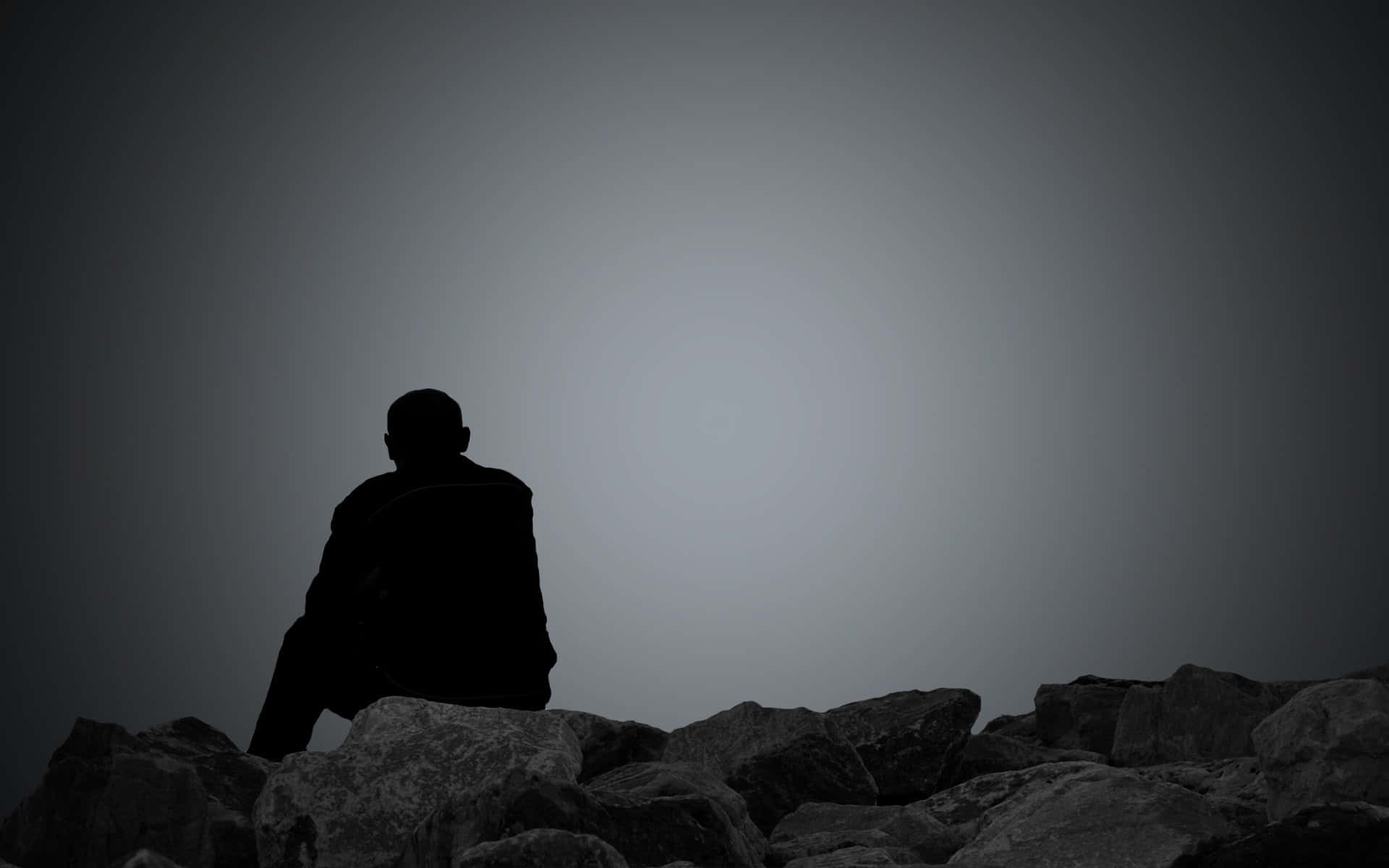 Depressing Profile 桌布 壁紙 — 一個孤獨的剪影坐在參差不齊的岩石上，背景是漸變的灰色天空，喚起孤獨和靜默的沉思。