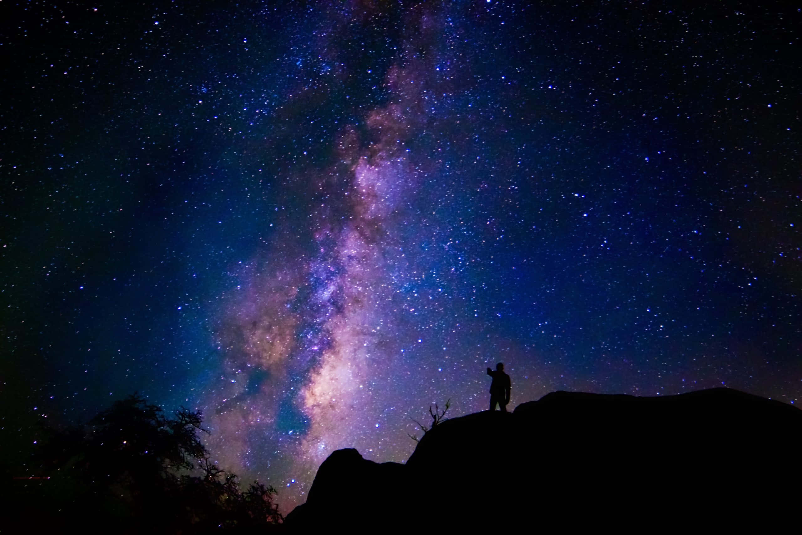 夜空 桌布 壁紙 — 一個孤獨的剪影站在岩石山脊上，仰望充滿銀河系鮮豔紫色和藍色色調的壯麗星空。