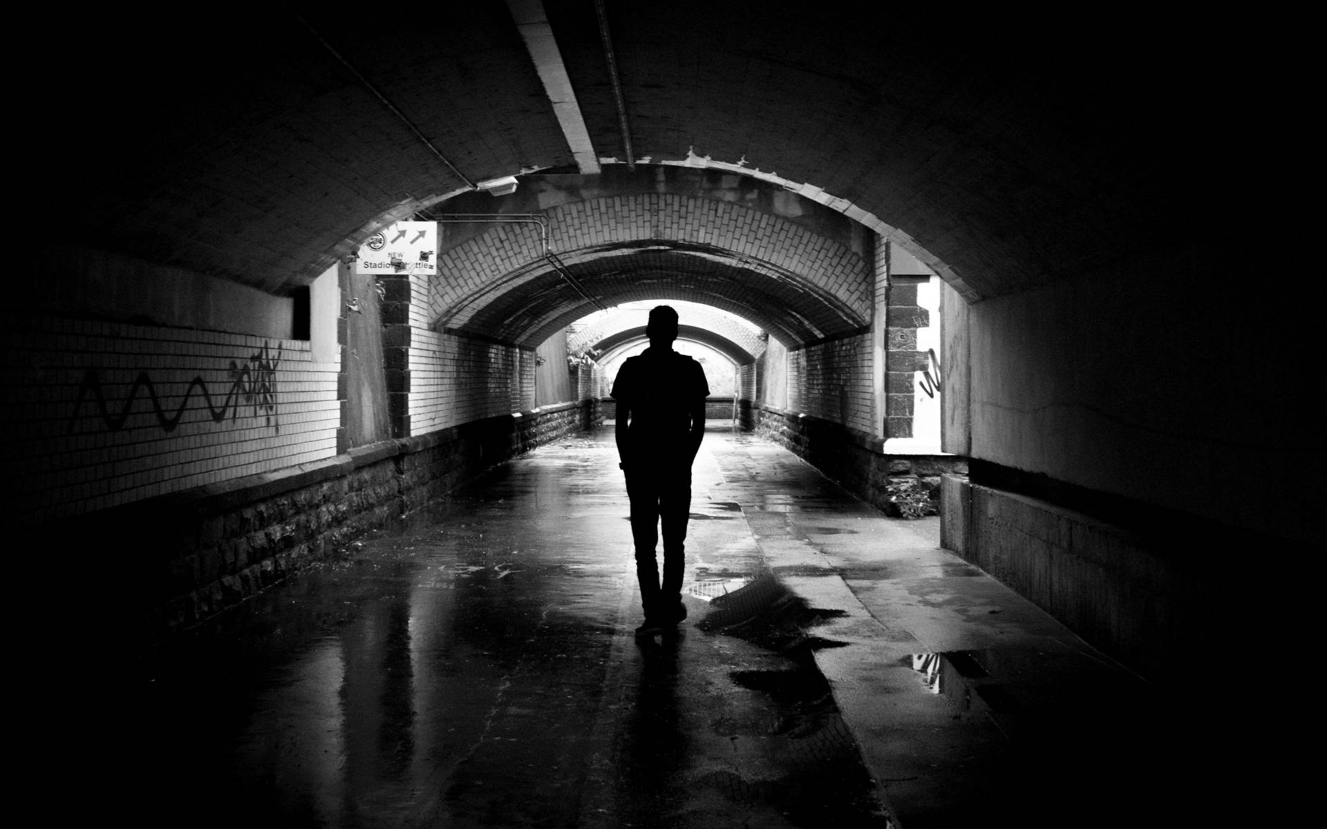 Alone 桌布 壁紙 — 一個孤獨的剪影走在一條潮濕、拱形的磚砌隧道中，隧道上有塗鴉，以強烈的黑白對比呈現。