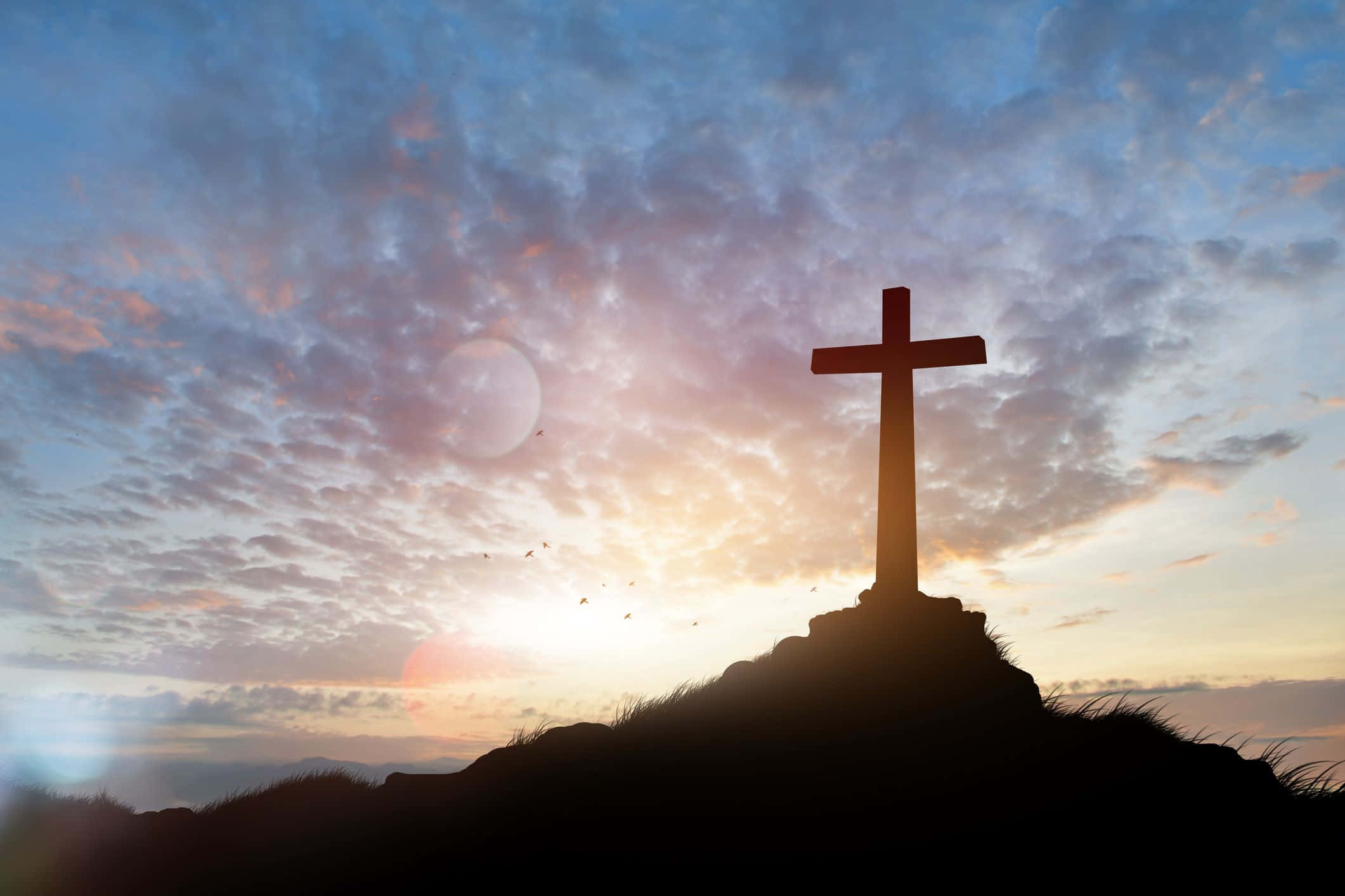 Religious Easter 桌布 壁紙 — 一個孤獨的十字架剪影立於山丘頂上，背景是戲劇性的日出天空，散落著雲朵和鏡頭光暈。