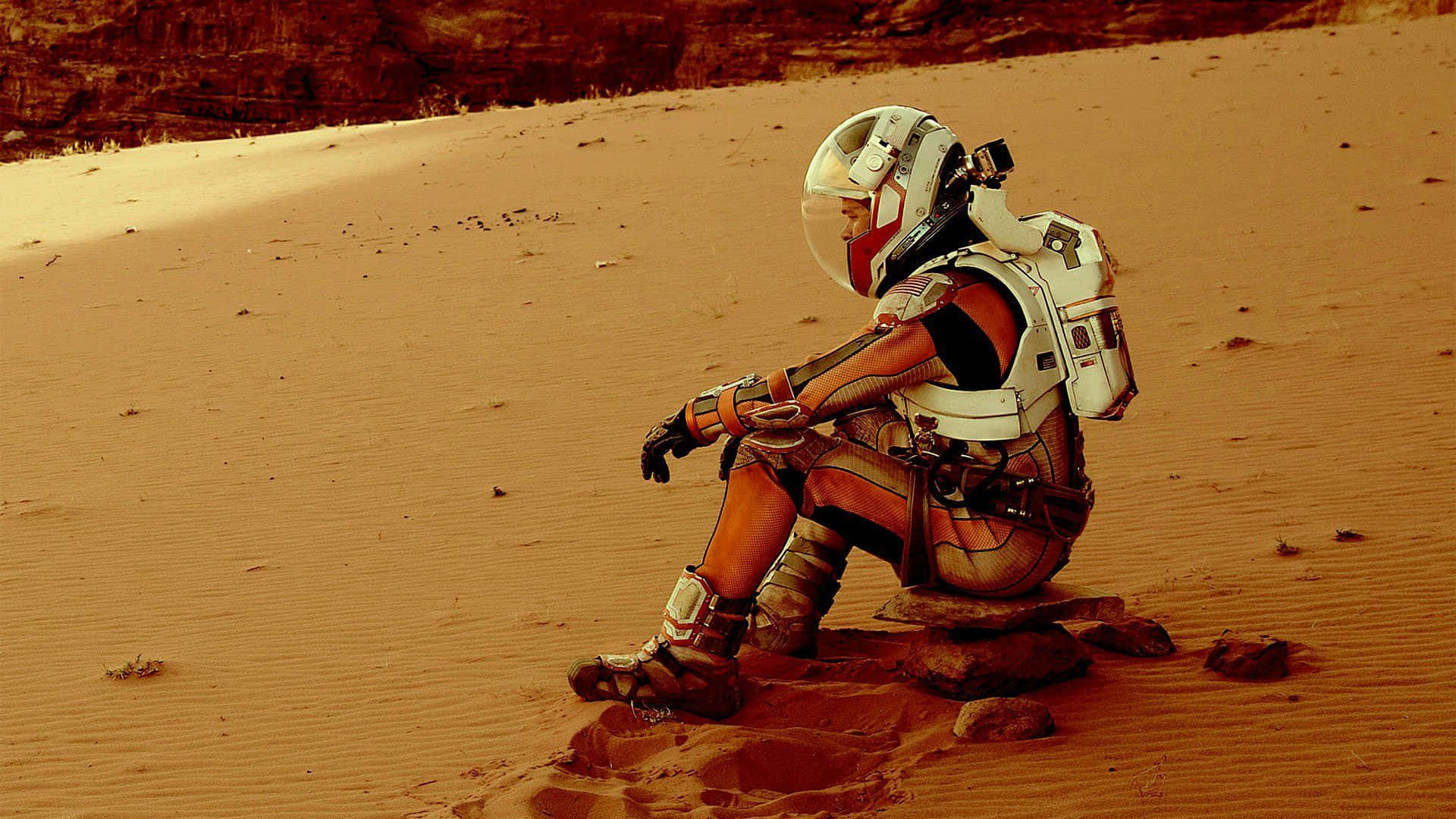 The Martian 桌布 壁紙 — 一個孤獨的太空人戴著白色頭盔，在溫暖的陽光下，沉思地坐在廣闊的微紅色沙漠中的一塊岩石上。
