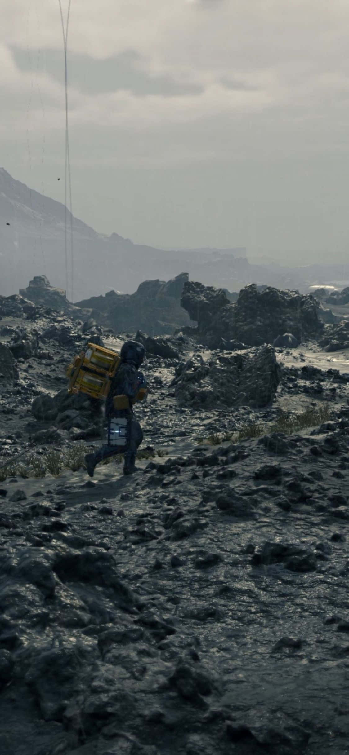 iPhone X Death Stranding 桌布 壁紙 — 一個孤獨的太空人穿著笨重的太空衣，在朦朧的天空下，穿越一片荒涼多岩石的外星景觀，搬運一個黃色板條箱。