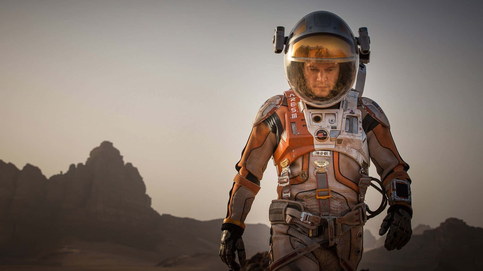 The Martian 桌布 壁紙 — 一個孤獨的太空人穿著細節豐富的橙色和灰色太空衣，站在一個荒蕪的外星行星上，岩石地形在朦朧的天空下。