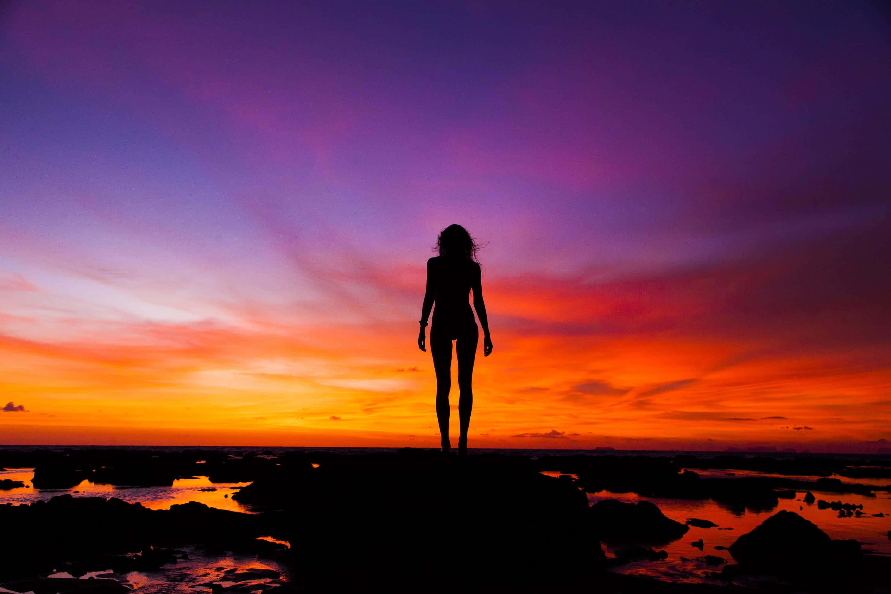 Sunset Silhouette 桌布 壁紙 — 一個孤獨的女人剪影站在岩石海岸上，下方是火燒般的日落天空，塗上鮮豔的橙色、紫色和粉紅色調。